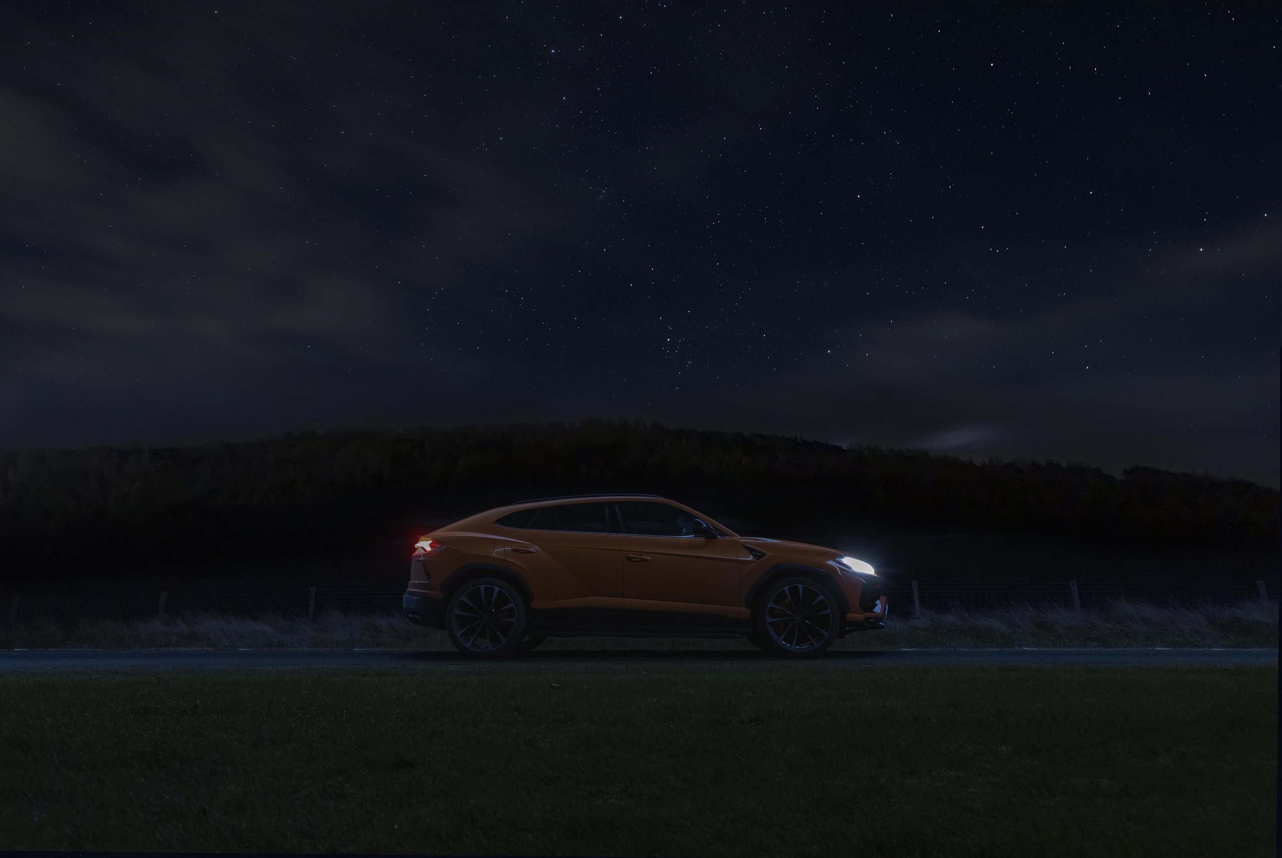 Lamborghini Sent Its Entire Lineup Into The Scottish Highlands, And The Real Destination Was The Stars 9 An orange lamborghini urus drives on a dark rural road under a starry night sky with headlights illuminated