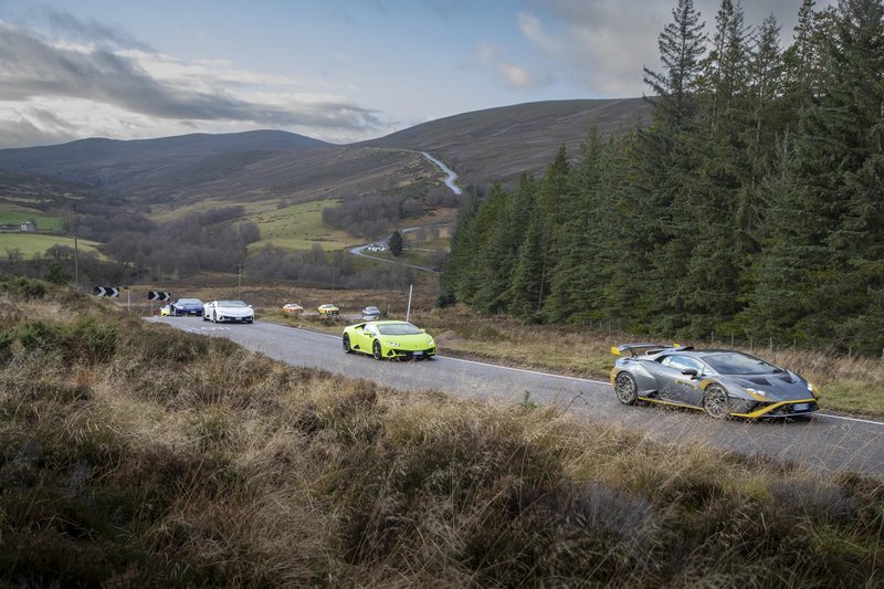 A convoy of Lamborghini supercars including a Huracán STO and Huracán EVO winds through a rugged Scottish Highland mountain road under cloudy skies