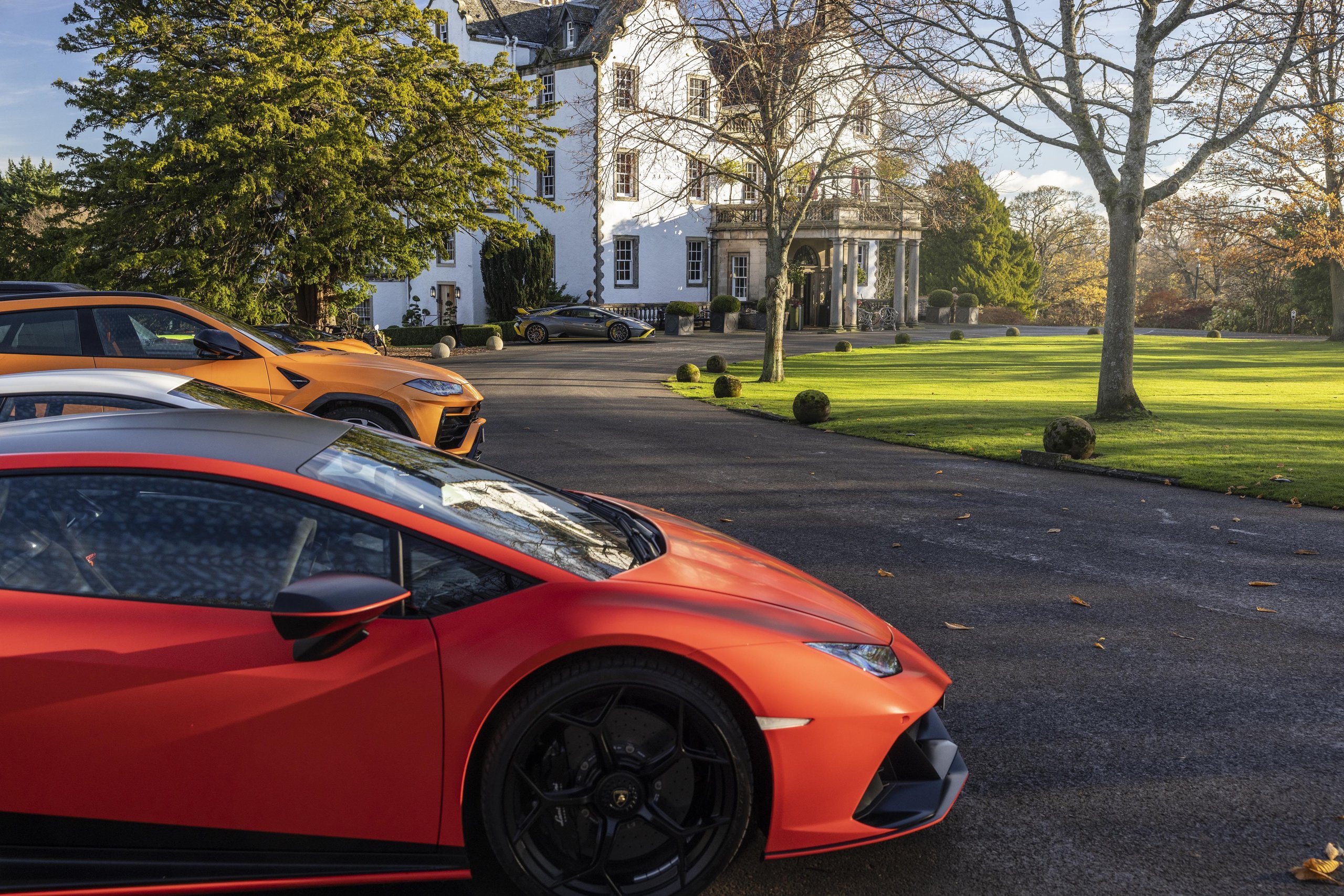 Lamborghini Sent Its Entire Lineup Into The Scottish Highlands, And The Real Destination Was The Stars 11 A red lamborghini huracán and orange urus parked on a paved driveway in front of a grand white scottish manor house surrounded by green lawns