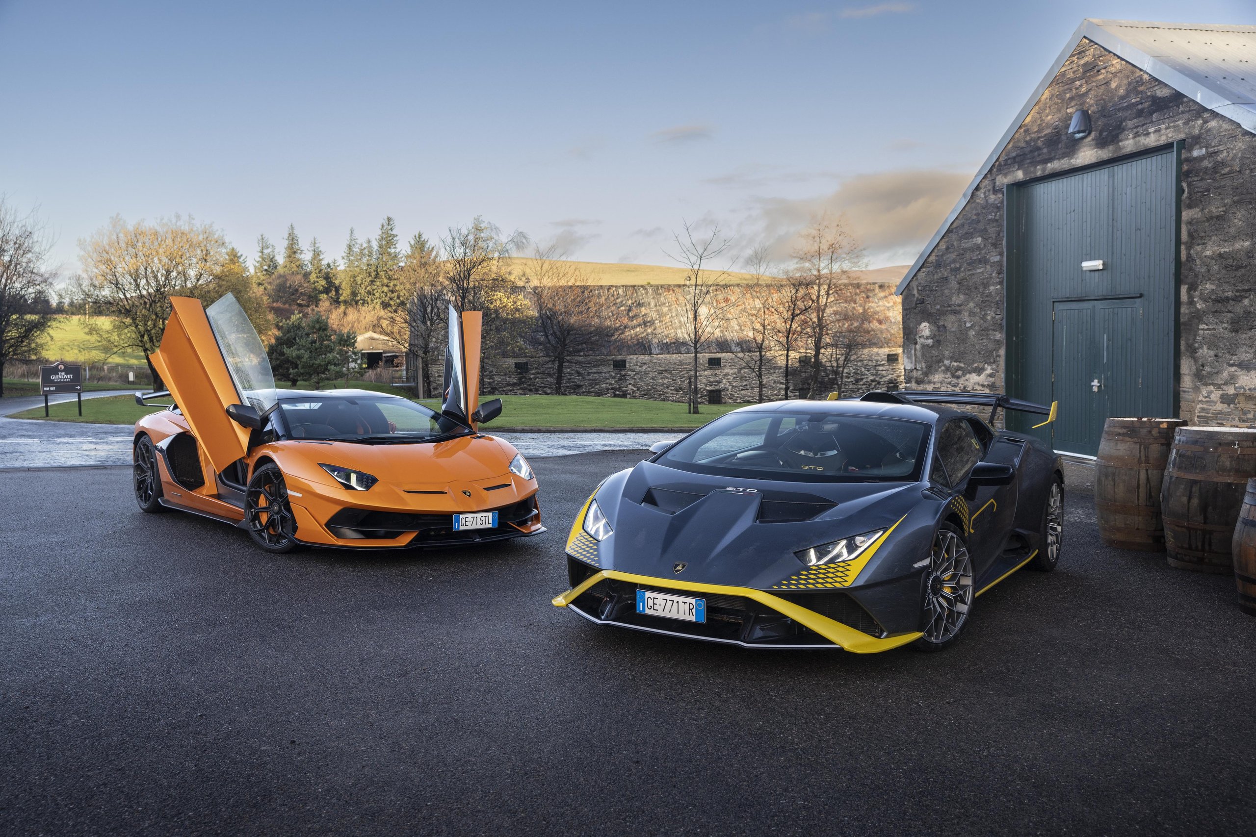 Lamborghini Sent Its Entire Lineup Into The Scottish Highlands, And The Real Destination Was The Stars 5 An orange aventador svj with scissor doors open parked beside a grey and yellow huracán sto in front of a rustic stone building with wooden barrels