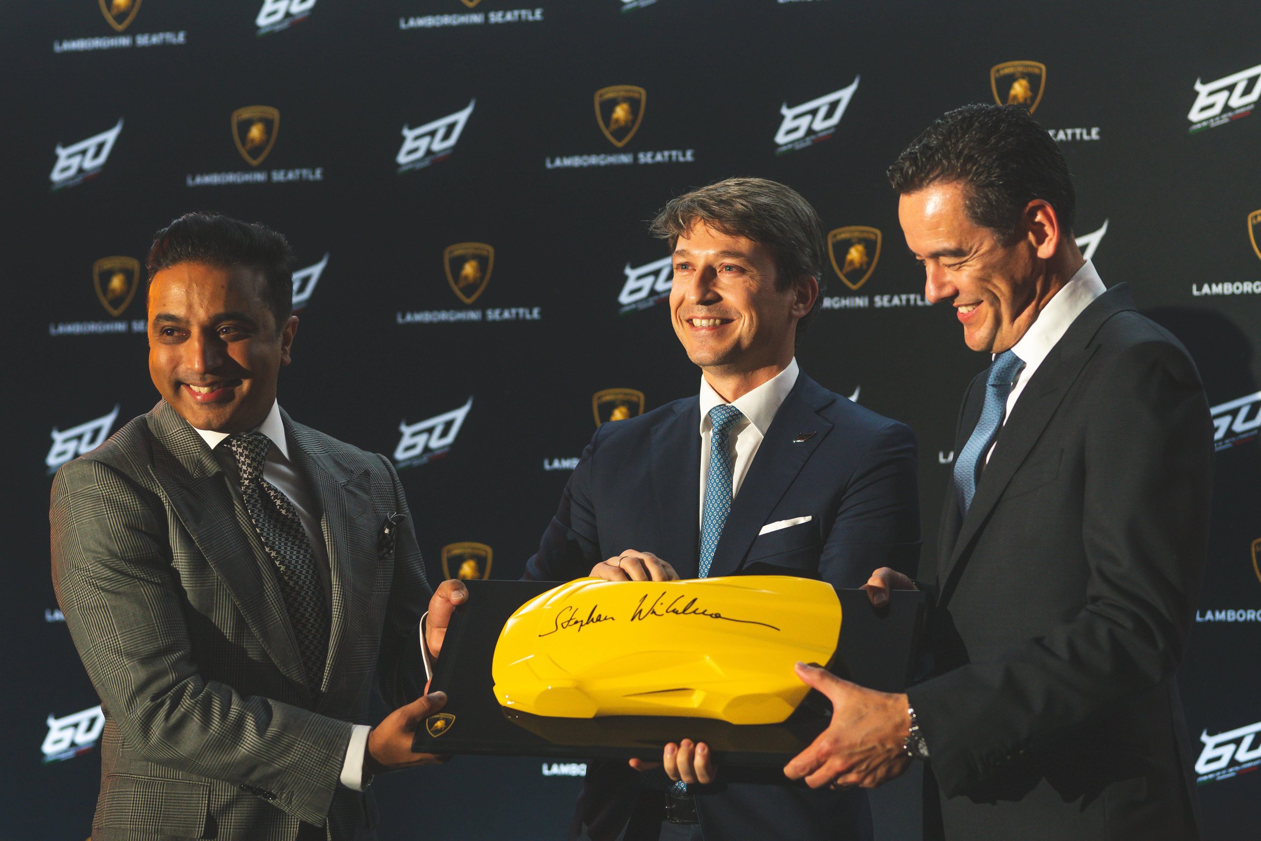 Lamborghini Seattle Opens In Bellevue With The Revuelto As Its Centerpiece, And That Pairing Tells You Everything About The Brand'S Strategy 7 Three men in suits holding a yellow lamborghini scale model at the lamborghini seattle grand opening with 60th anniversary logos