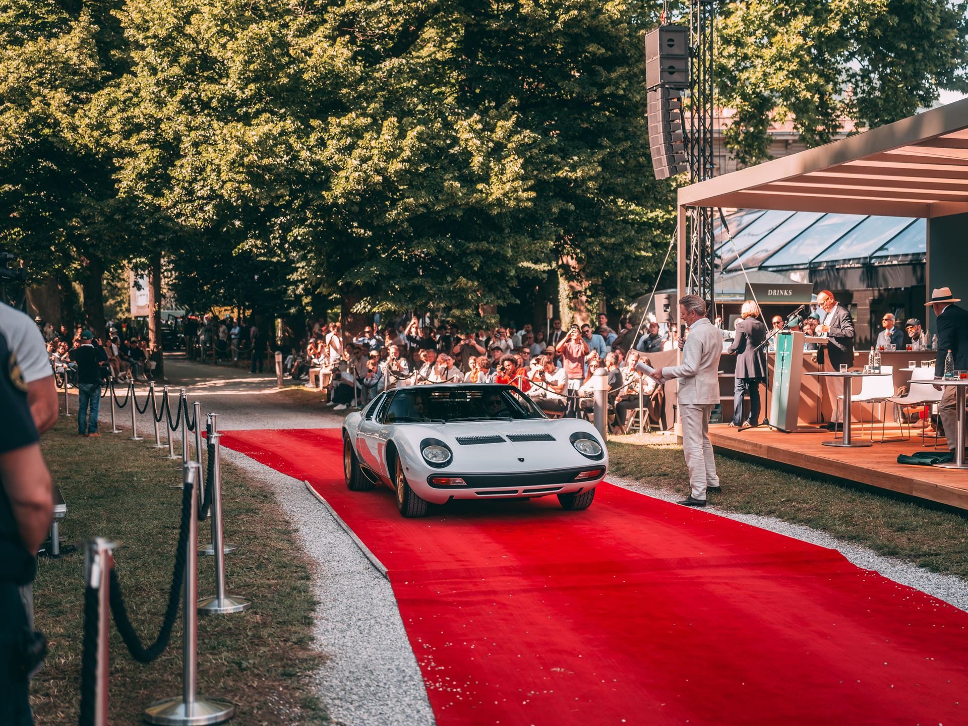 A white lamborghini miura sv presented on a red carpet at an exclusive outdoor event with spectators