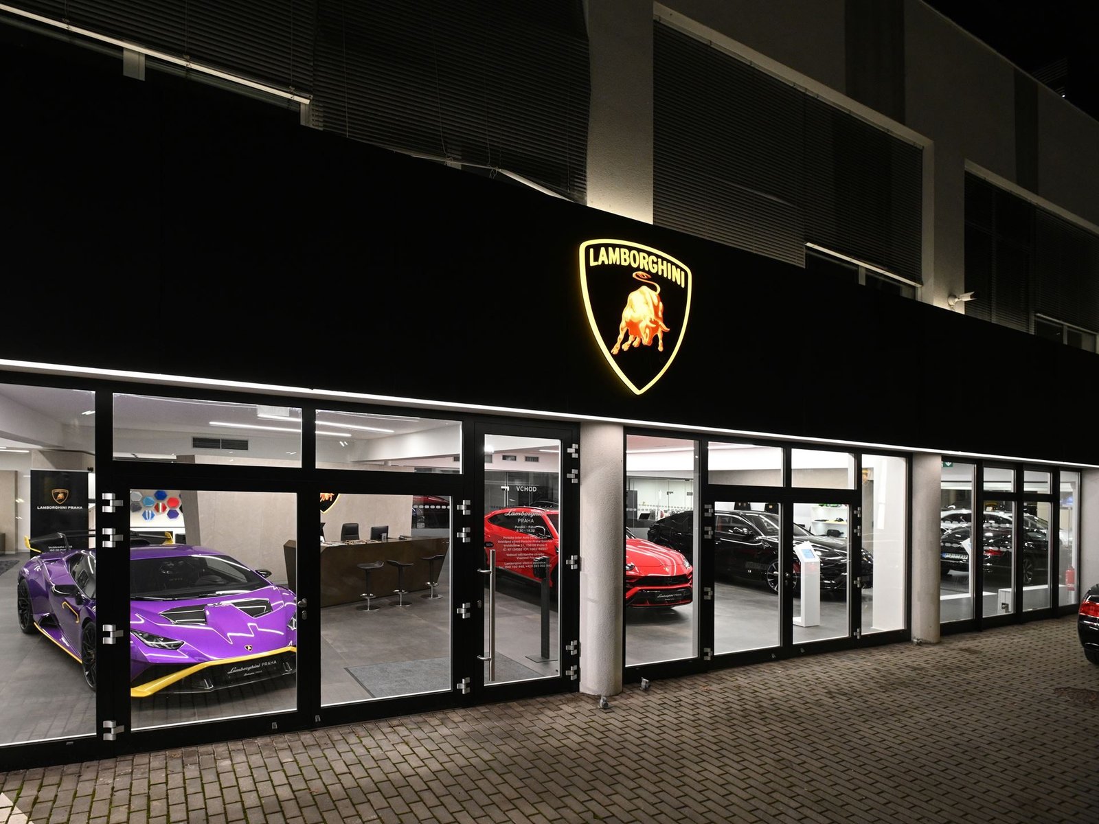 Exterior of the Lamborghini Prague showroom at night, with a purple Huracan Tecnica and red Urus visible through the glass beneath an illuminated Lamborghini logo