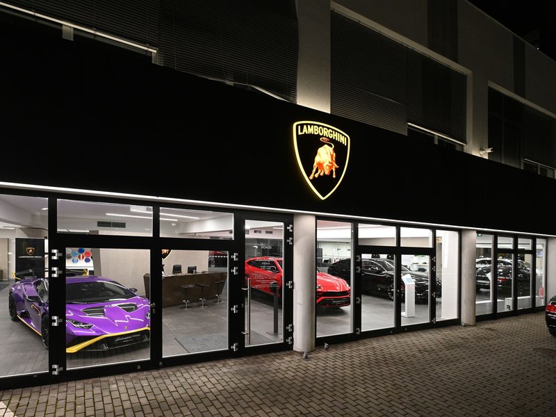 Exterior of the Lamborghini Prague showroom at night, with a purple Huracan Tecnica and red Urus visible through the glass beneath an illuminated Lamborghini logo