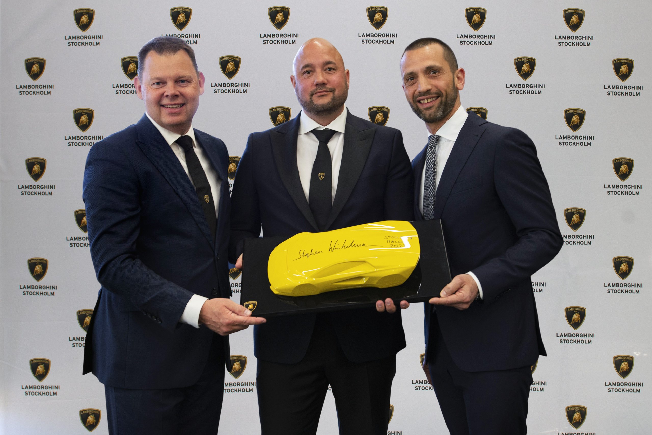 Three executives holding a yellow Lamborghini sculpture at the official inauguration event against a backdrop of Lamborghini Stockholm branding