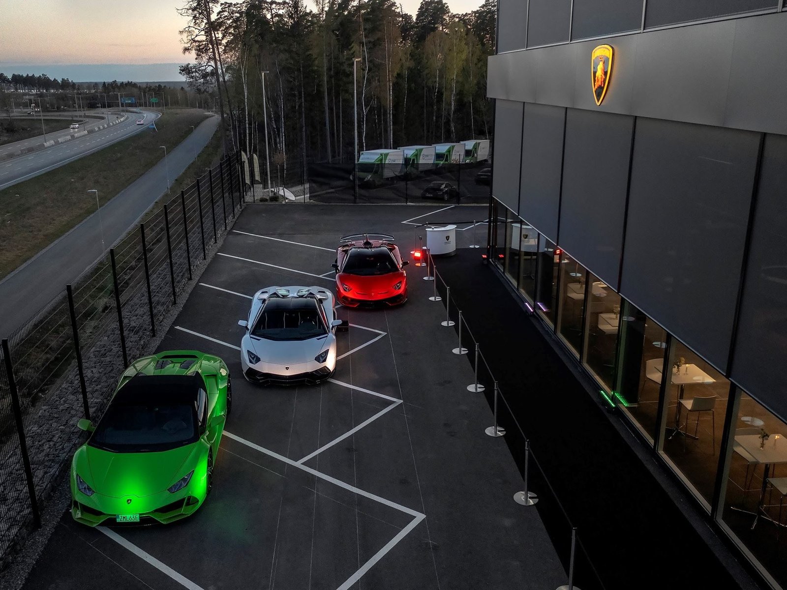 Aerial dusk view of the new Lamborghini Stockholm showroom with a Huracán, Aventador, and Urus parked outside the modern facility along highway E4
