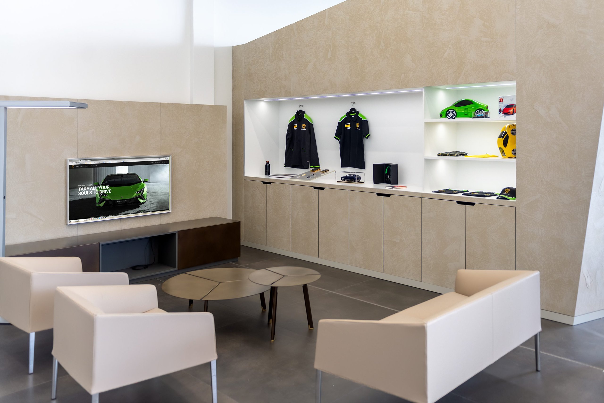 Comfortable lounge area inside the Lamborghini Stockholm showroom with modern seating, a display screen, and branded merchandise shelves