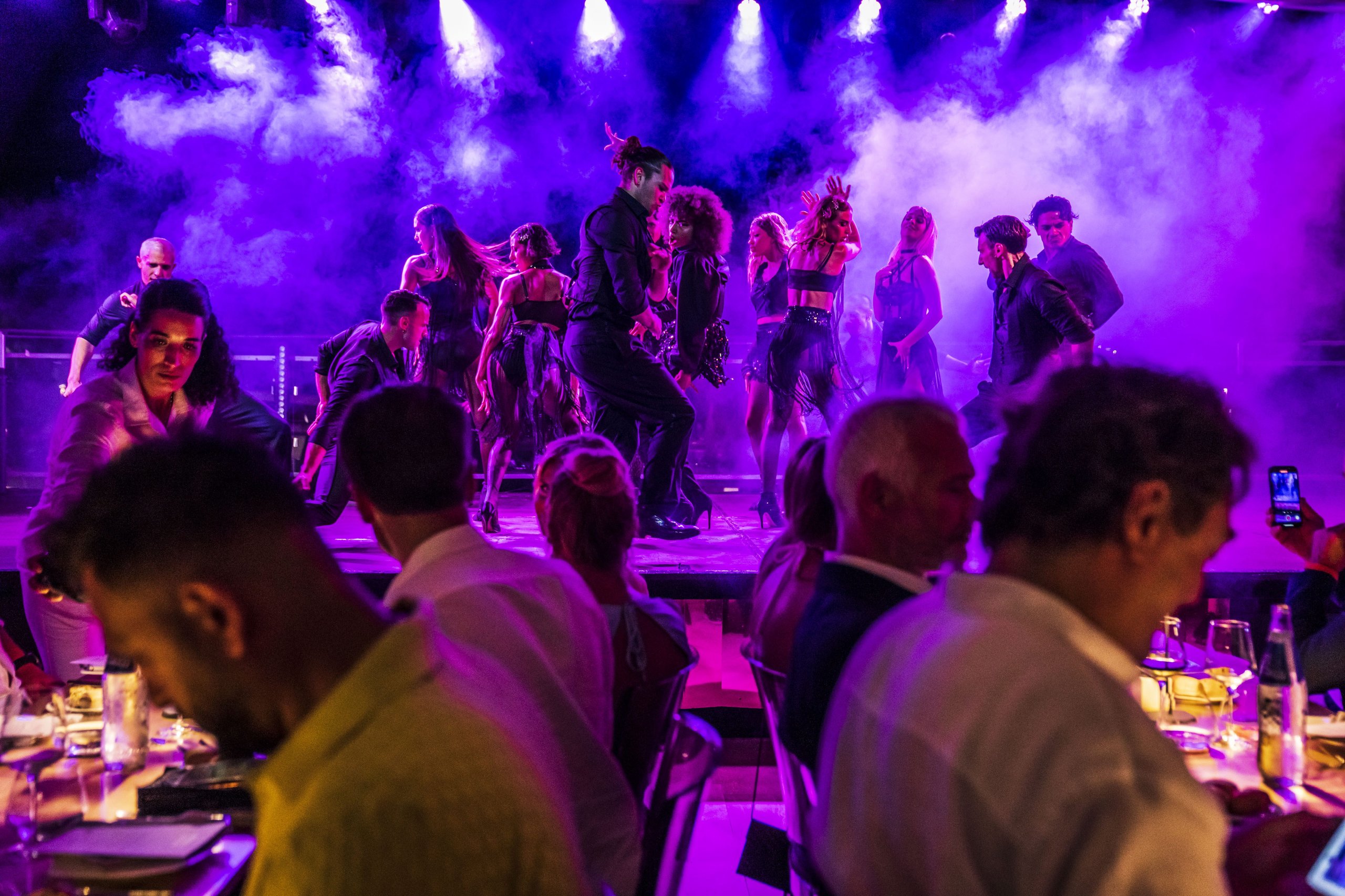 Dancers perform on stage under purple lighting at the Lamborghini Summer Dinner, viewed from guests' tables