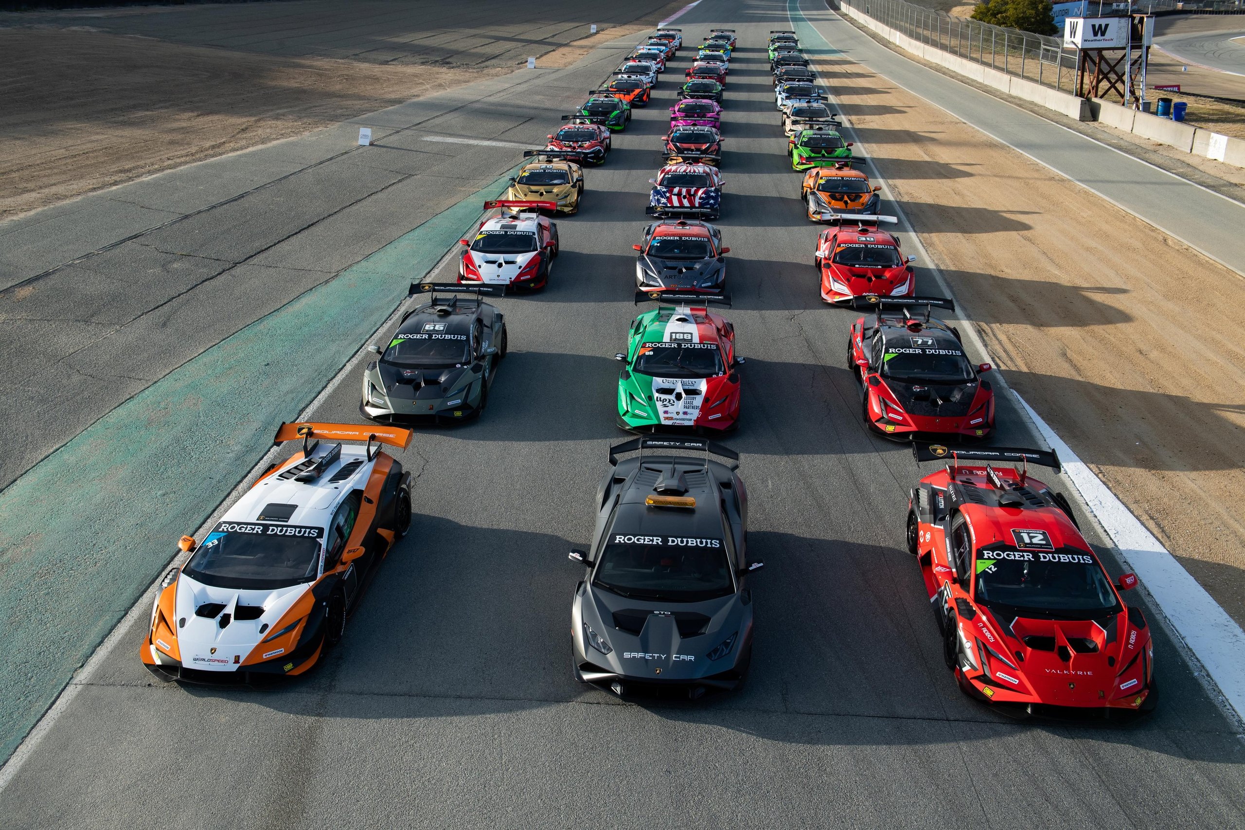 Lamborghini Super Trofeo'S 2024 Calendar Puts Customer Racing On The World'S Biggest Stages, Starting With Le Mans 7 Aerial view of numerous lamborghini huracán super trofeo evo2 race cars parked in formation on a racetrack with pit lane and grandstands visible