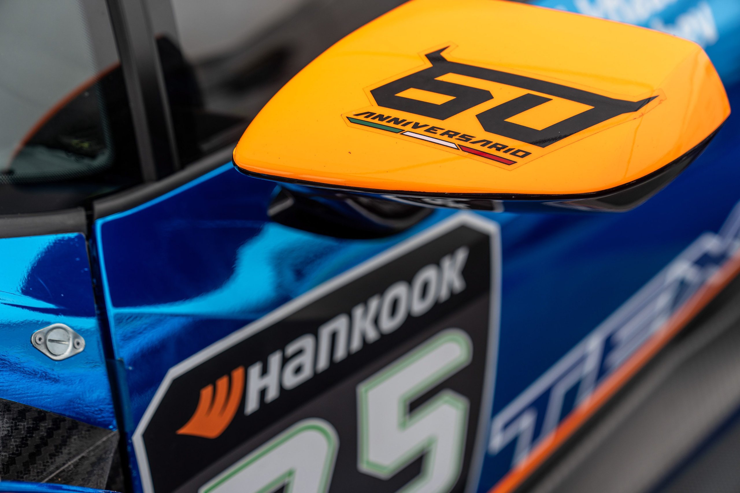 49 Huracán Evo2S At Spa: Lamborghini'S One-Make Series Celebrates 60 Years Where It All Began 5 Close-up of a huracán super trofeo evo2 side mirror with orange 60 anniversario logo against a blue livery and hankook decal