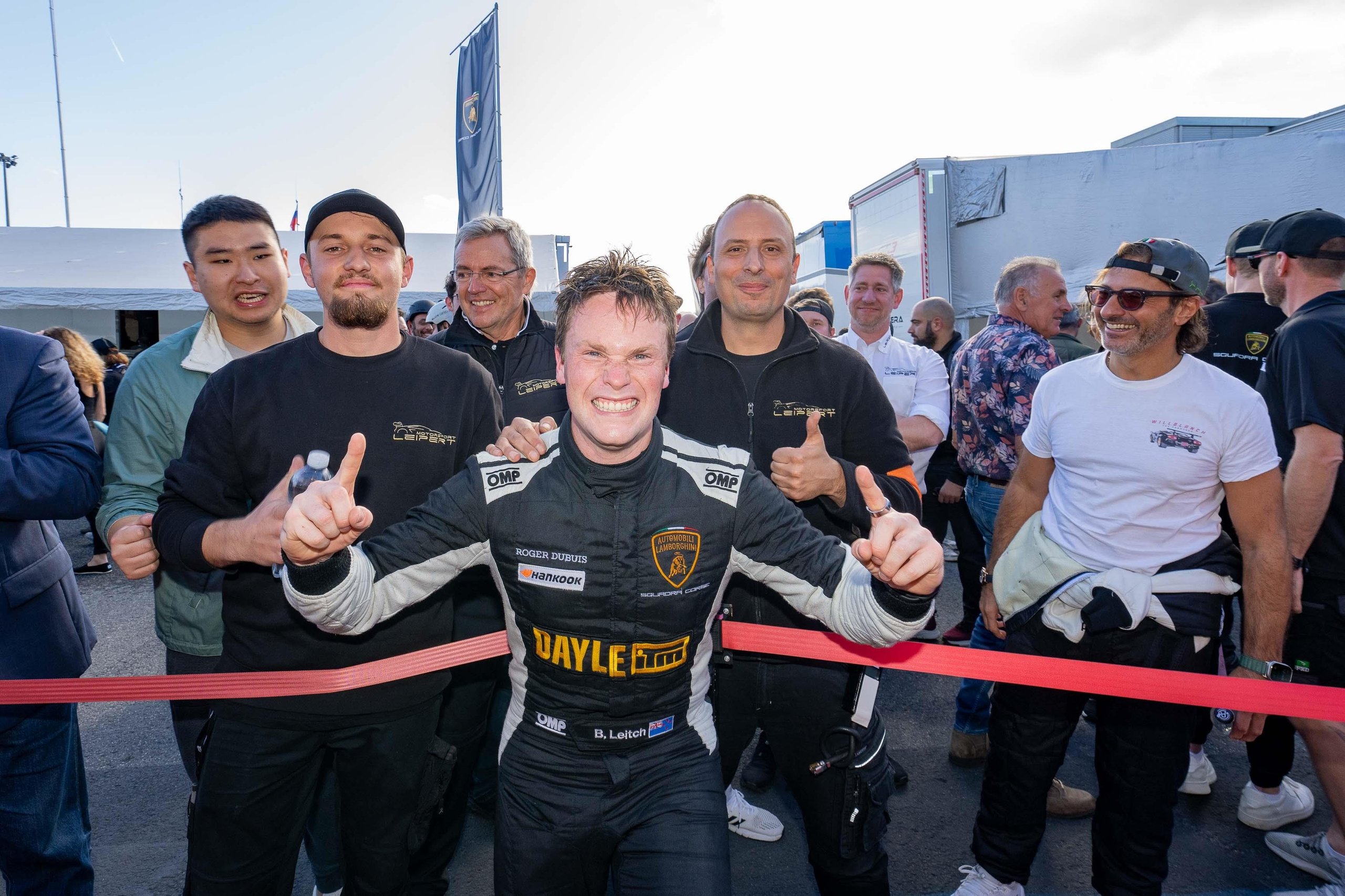 Leitch Claims European Pro Title In Thrilling Lamborghini Super Trofeo Finale That Crowned Champions Across Three Continents 7 Jubilant race car driver in racing suit giving two thumbs up surrounded by celebrating team members in the paddock