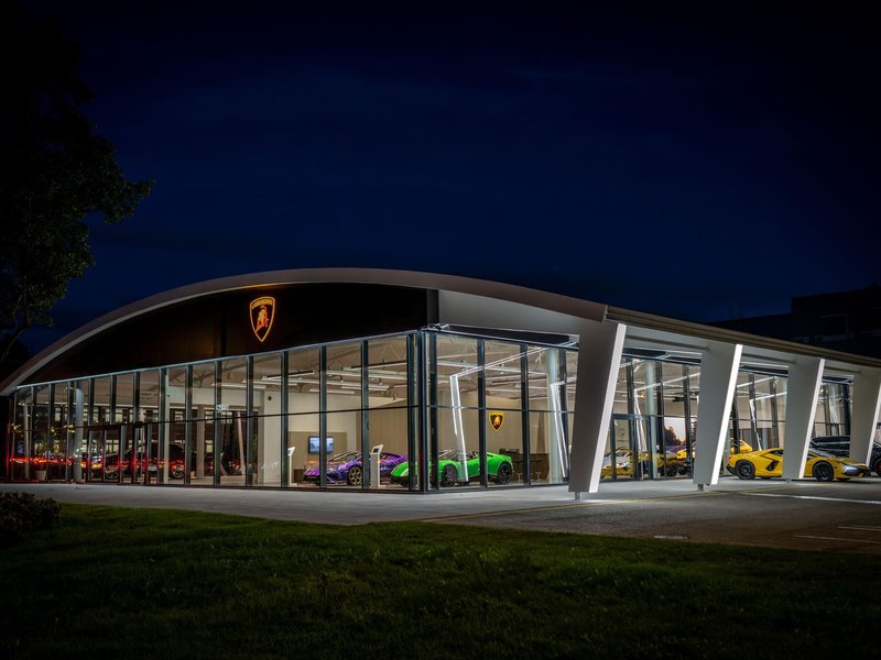 Exterior night shot of the new Lamborghini Tallinn showroom with brightly lit supercars visible through the glass facade