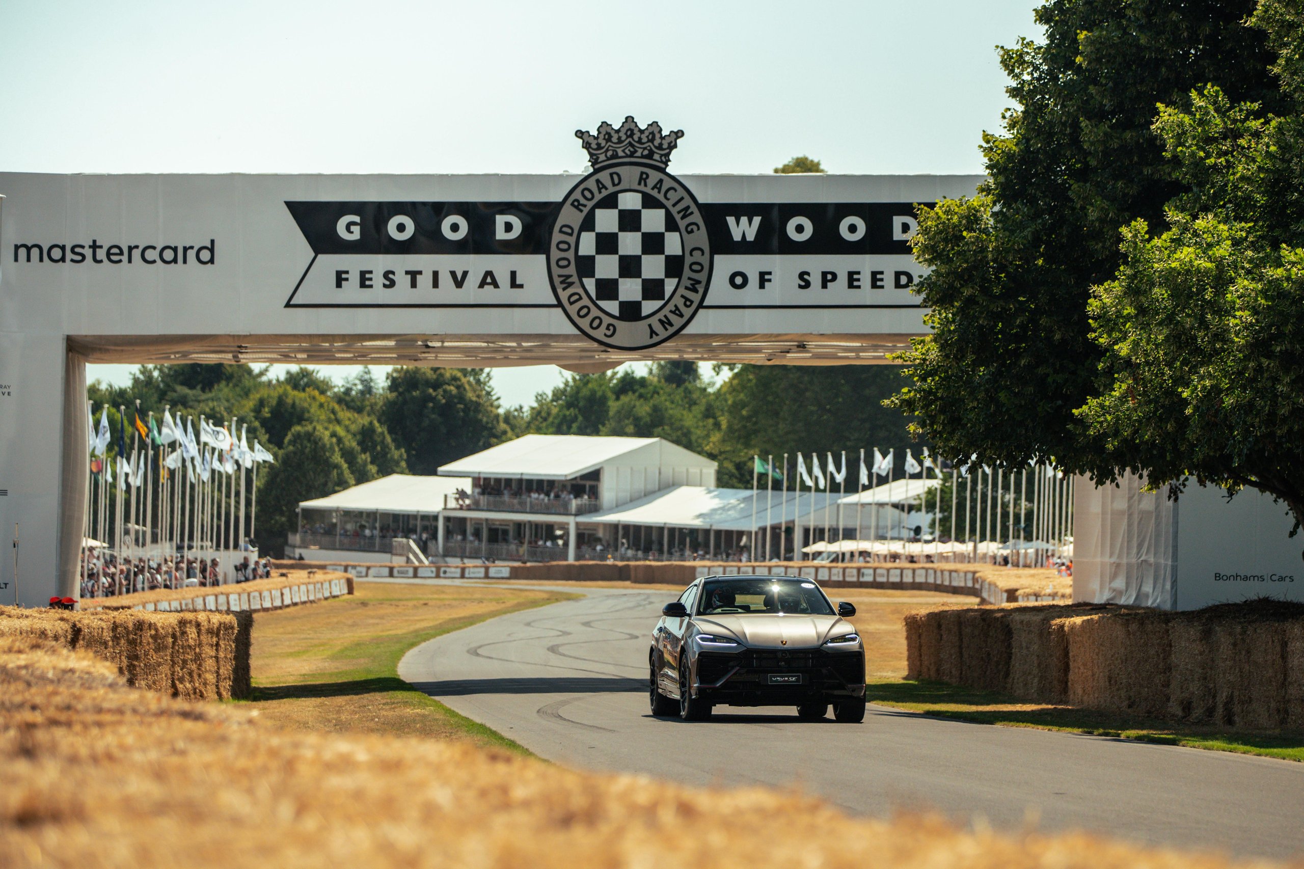 Grey lamborghini urus se driving on the goodwood festival of speed hill climb past hay bales and the event banner
