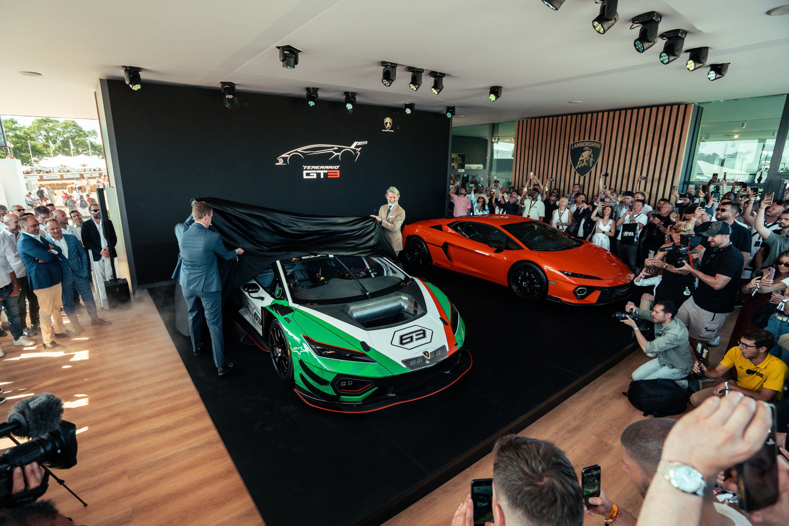 Two men unveil the lamborghini temerario gt3 from under a black cover, revealing its tricolore livery, with an orange revuelto and large audience visible