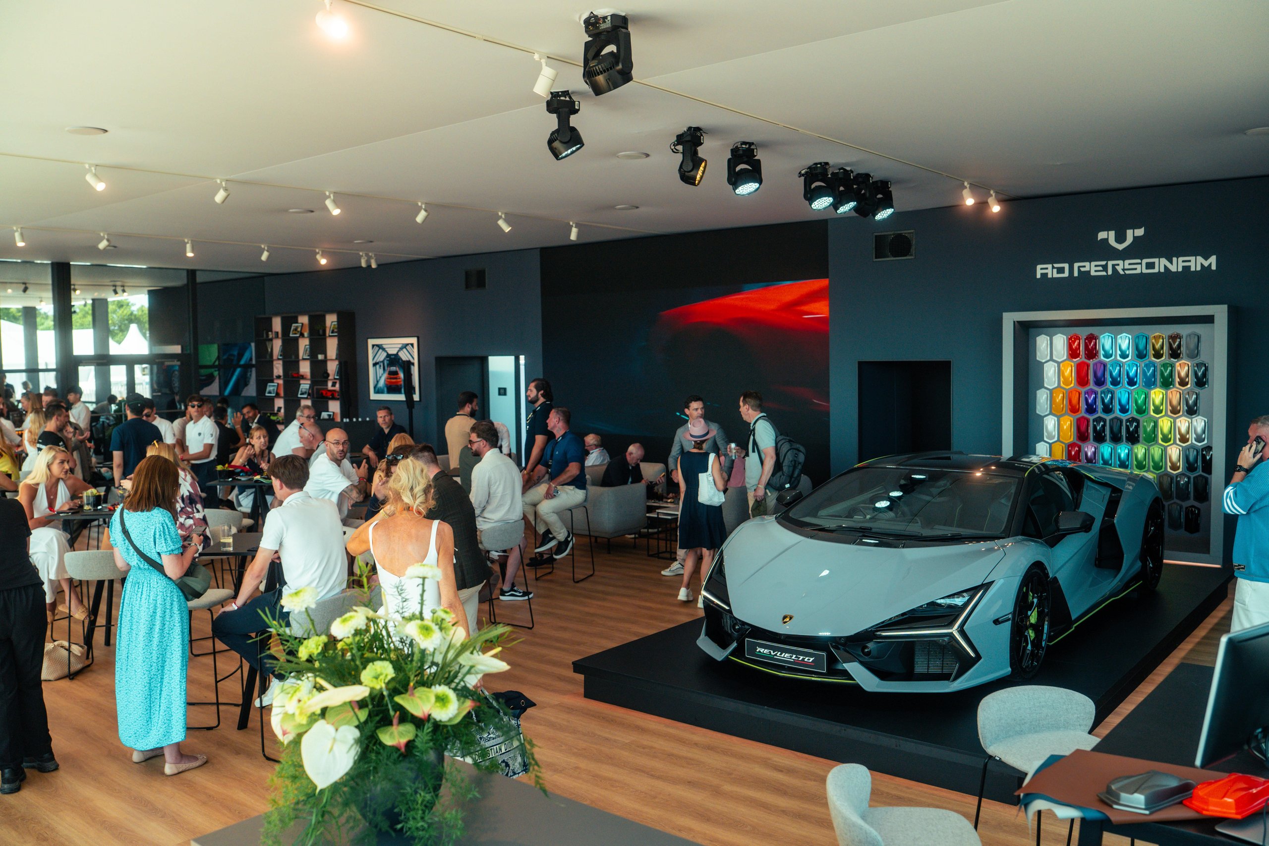 Grey lamborghini revuelto on a raised platform inside the lamborghini lounge, with guests and an ad personam customization display visible