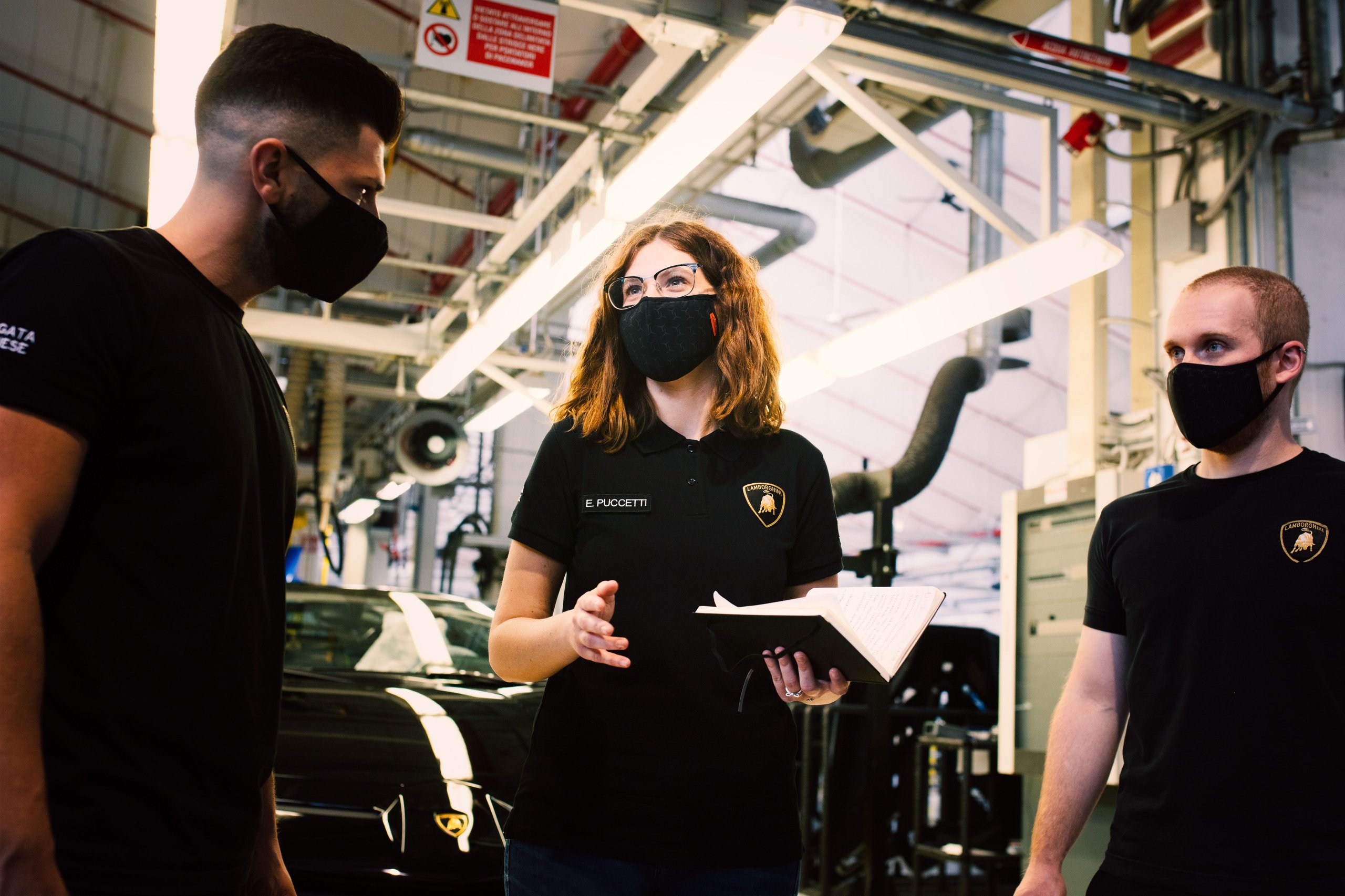 Three lamborghini employees discuss production on the factory floor with a black lamborghini huracan visible behind them