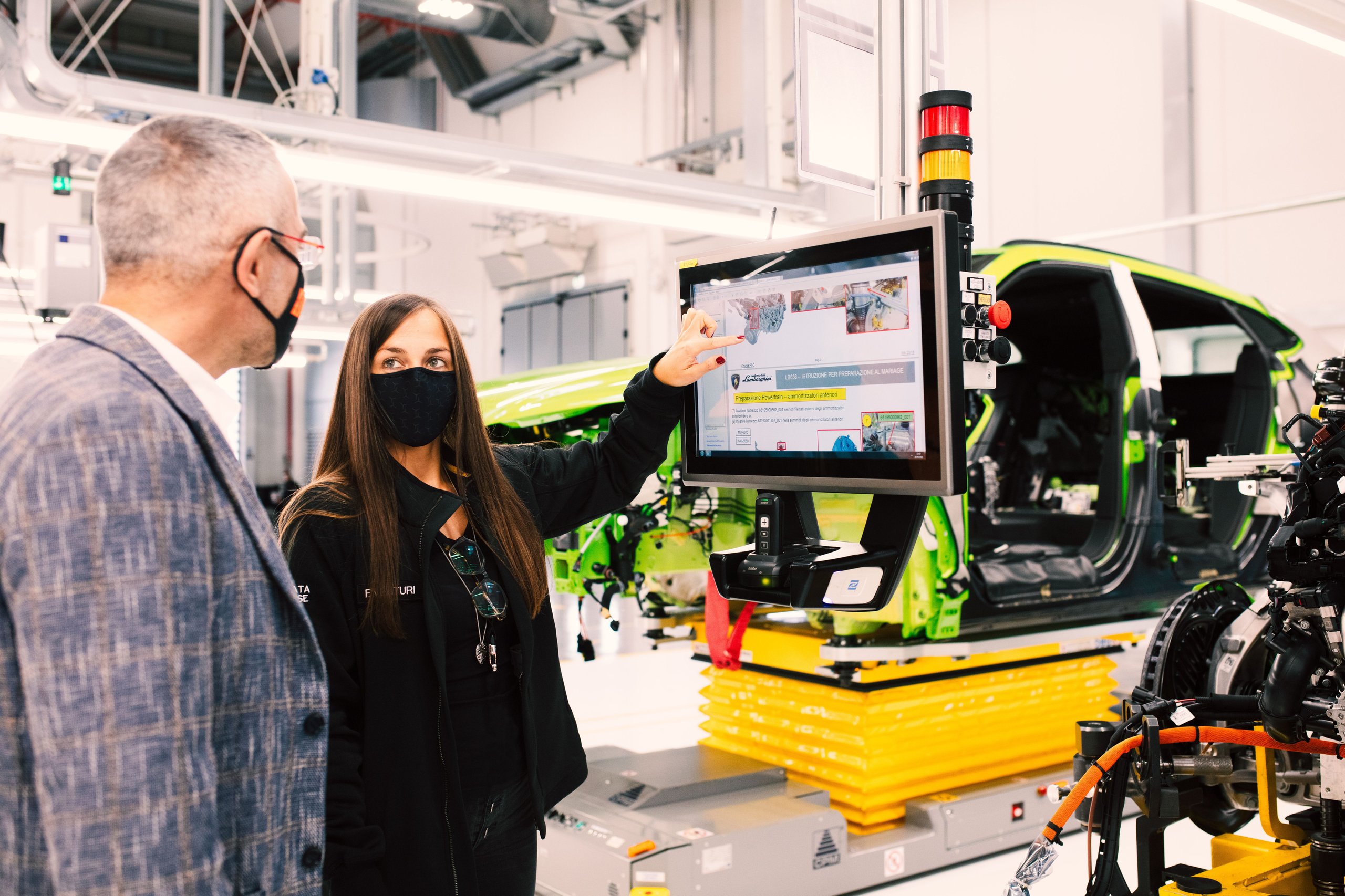 A female lamborghini employee points to a digital display while discussing with a colleague, with a partially assembled green urus in the background