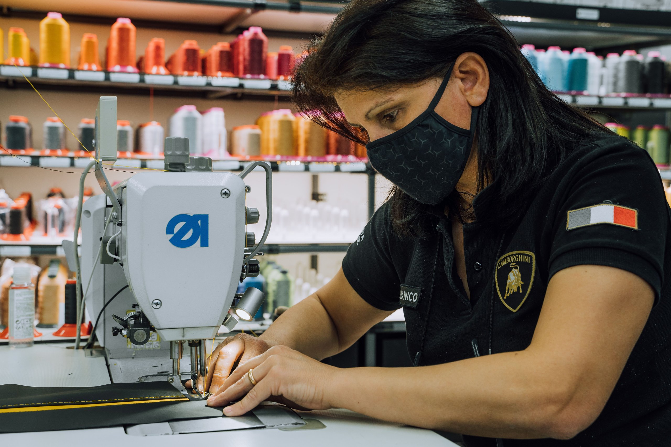 Close-up of a lamborghini employee operating a sewing machine to stitch black and yellow interior material, with spools of thread visible
