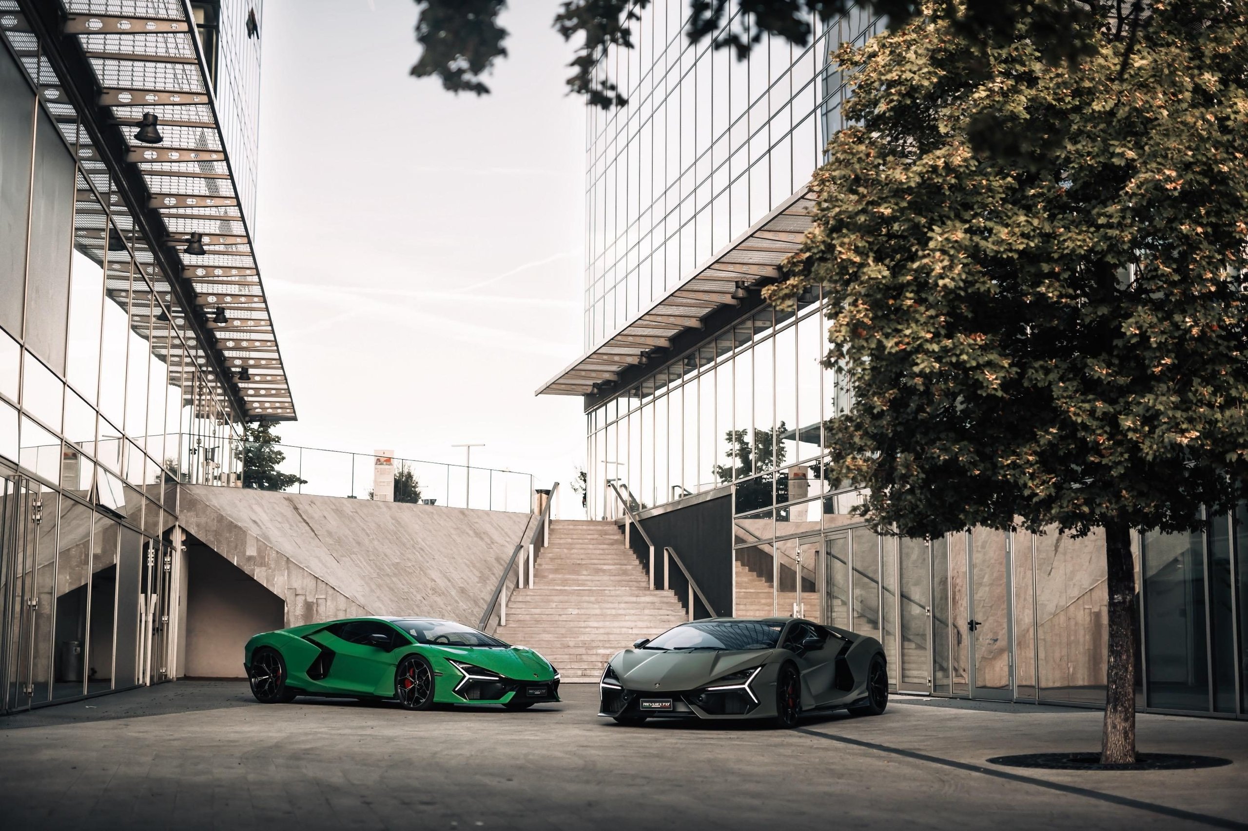 Two lamborghini revuelto models in bright green and matte grey parked in front of modern glass buildings