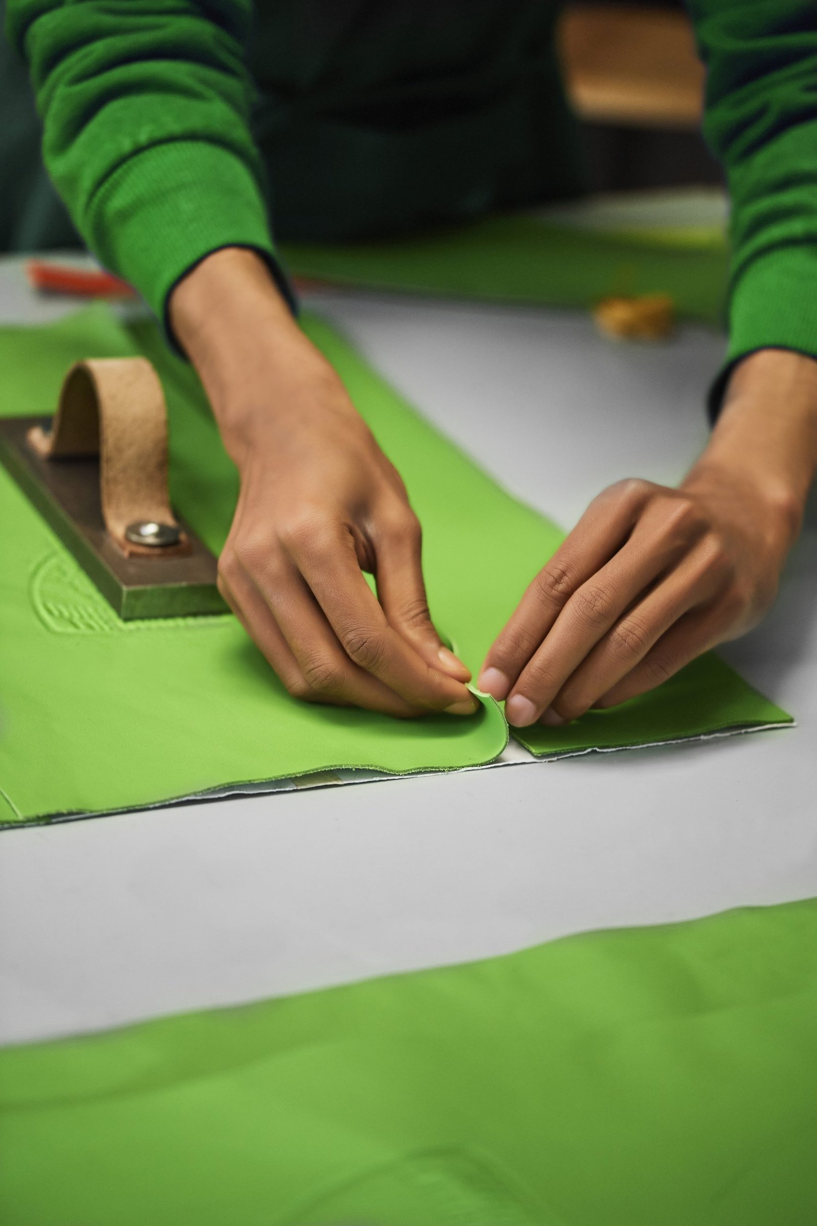 Artisan hands carefully folding green leather at Cartiera's workshop with a leather press visible in the background