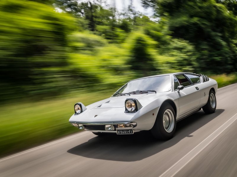 White Lamborghini Urraco P250 driving dynamically on a winding country road with blurred green trees in the background