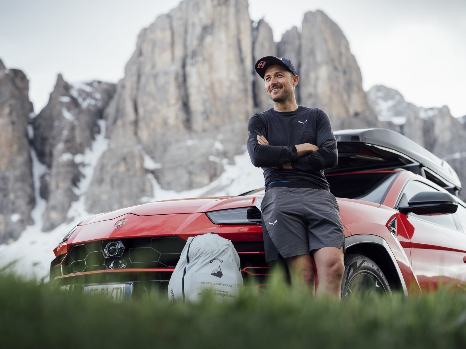 Aaron Durogati leaning against a red Lamborghini Urus with roof box in front of snow-capped Alpine mountains
