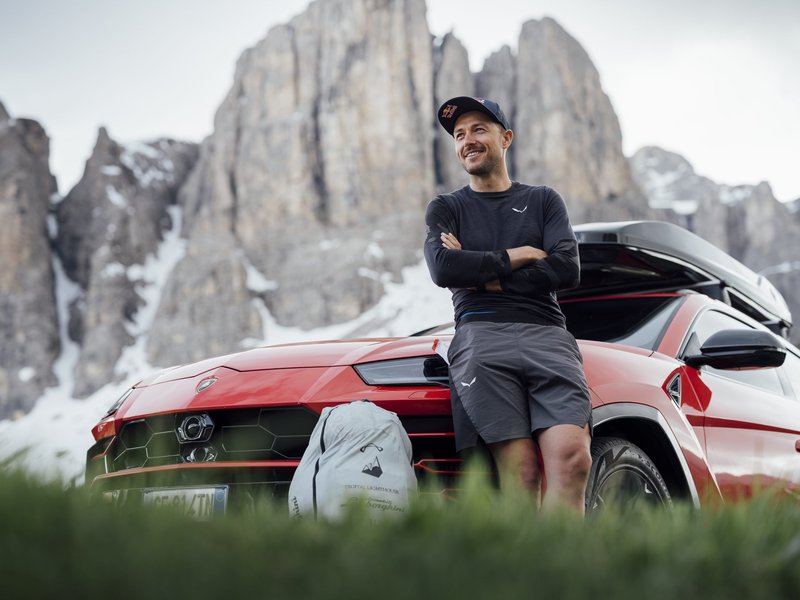 Aaron Durogati leaning against a red Lamborghini Urus with roof box in front of snow-capped Alpine mountains