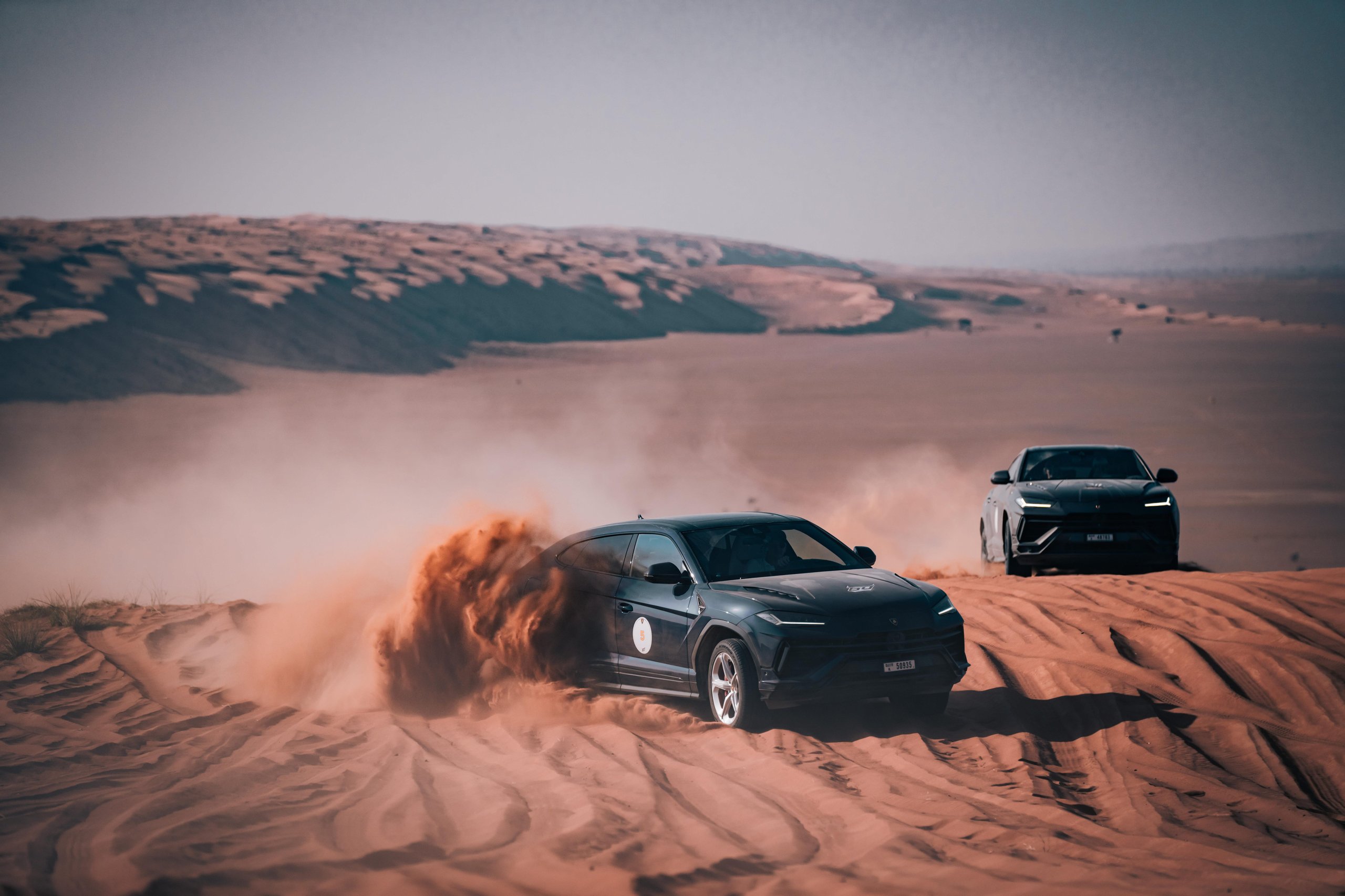 Black lamborghini urus s kicking up a large cloud of sand while driving aggressively through desert dunes