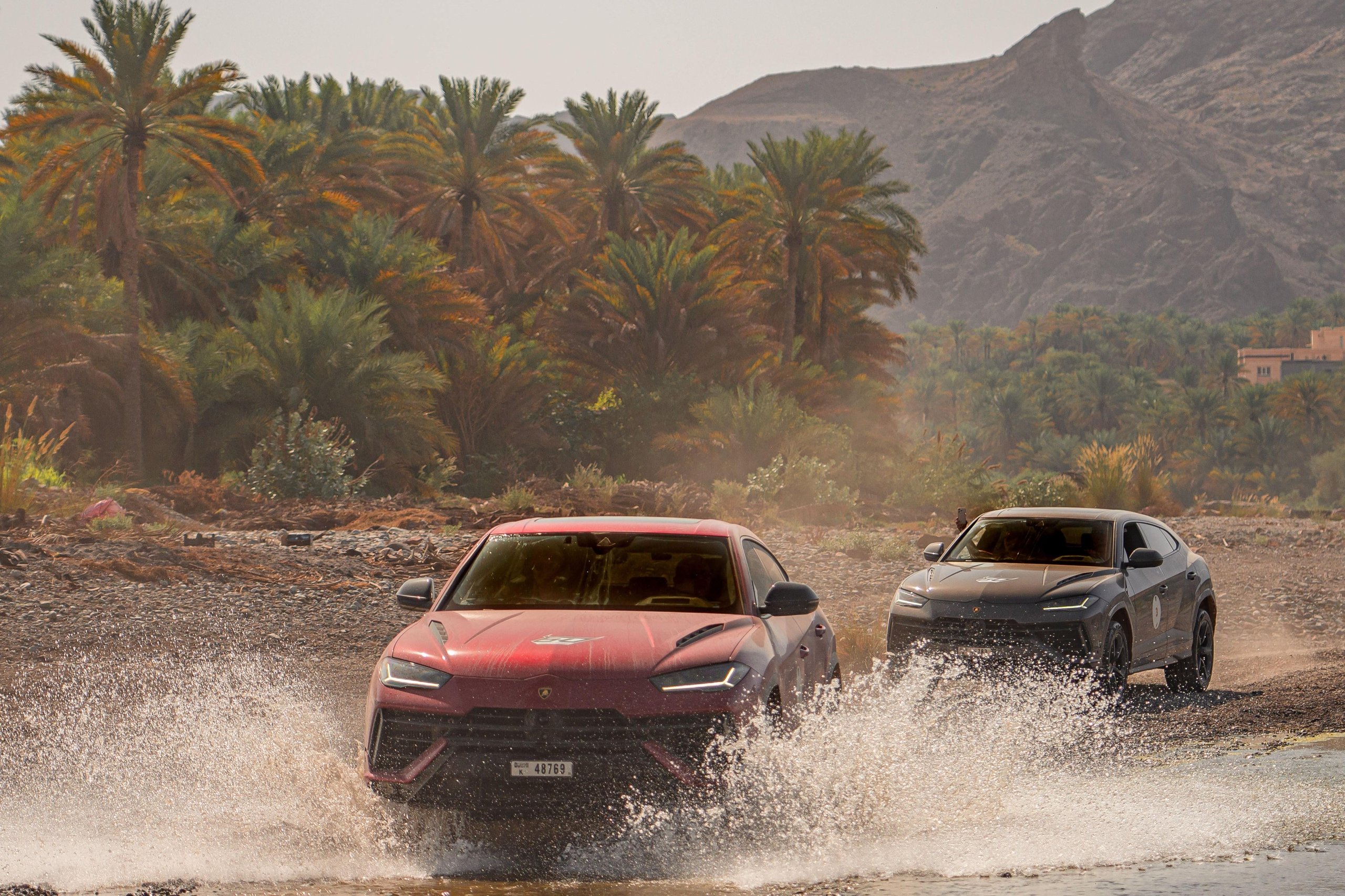 Red lamborghini urus s splashing through a shallow river crossing with palm trees and mountains in background