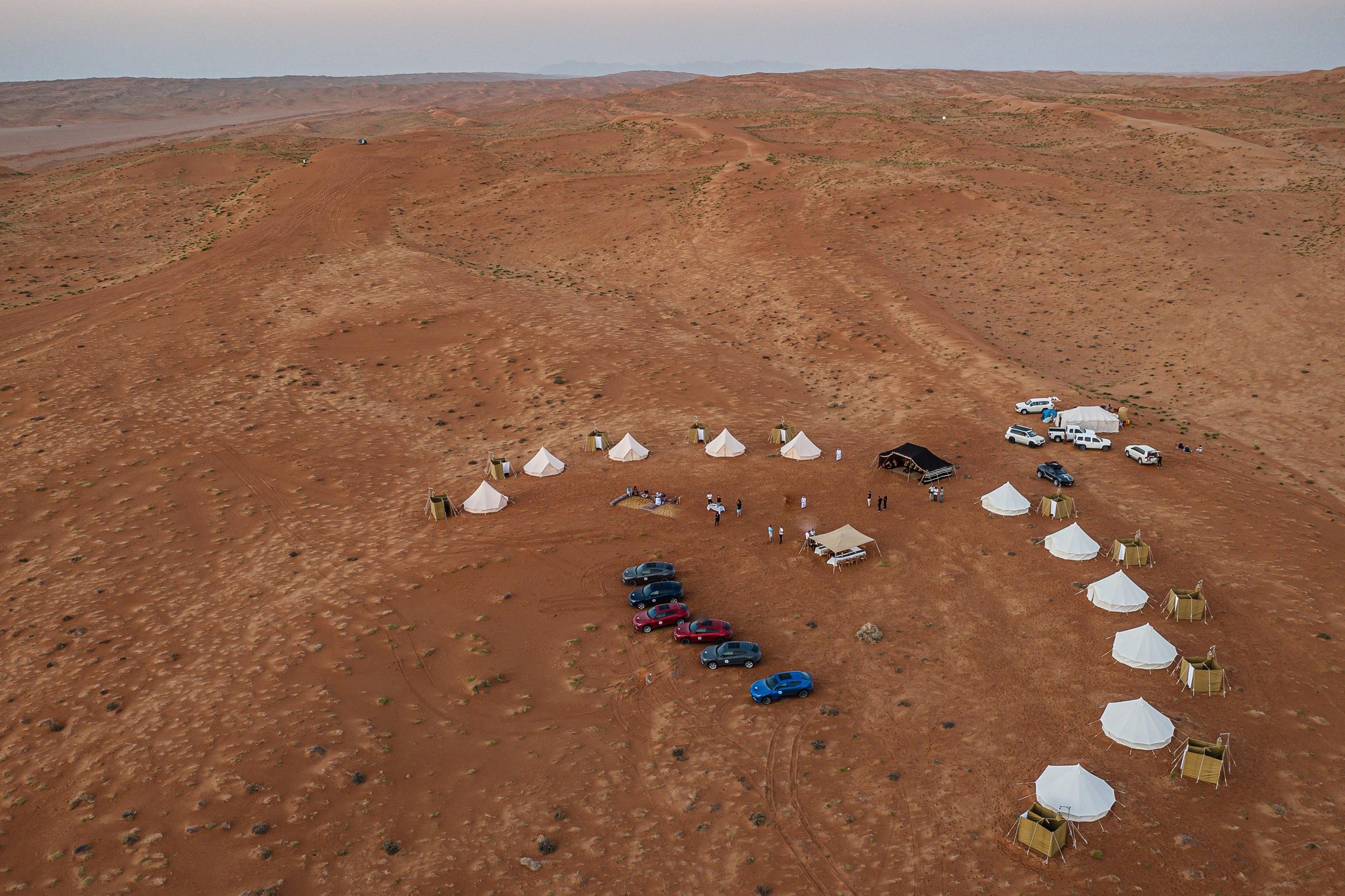 Aerial view of desert glamping camp with white tents arranged in a semi-circle on sand dunes, lamborghini urus vehicles parked nearby