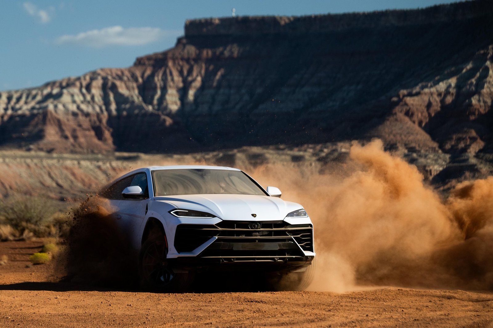 White Lamborghini Urus SE kicking up dust on a desert dirt track with rugged mountains in the background
