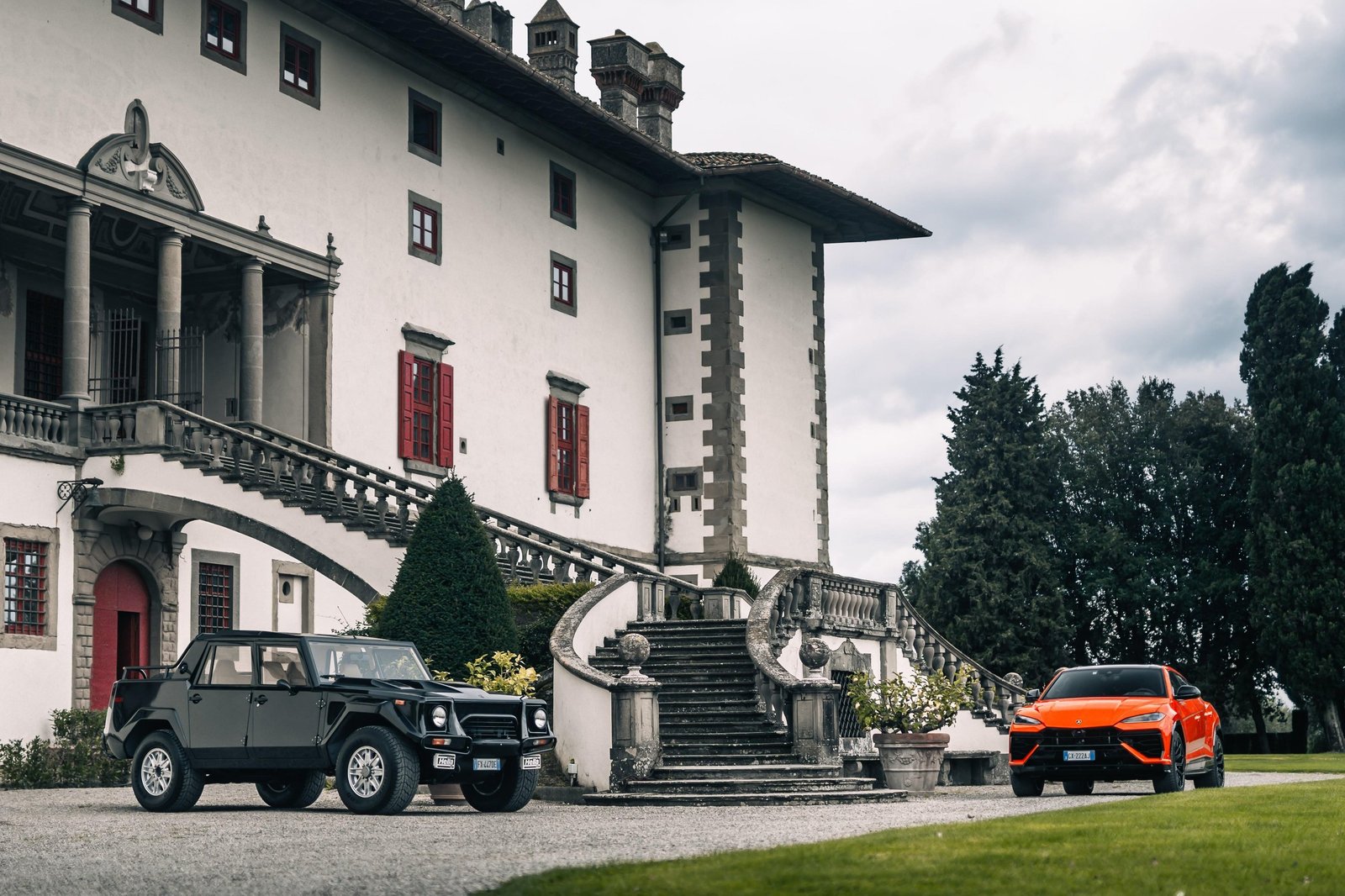 A black Lamborghini LM002 and an orange Lamborghini Urus parked together on a gravel driveway in front of a historic Italian villa