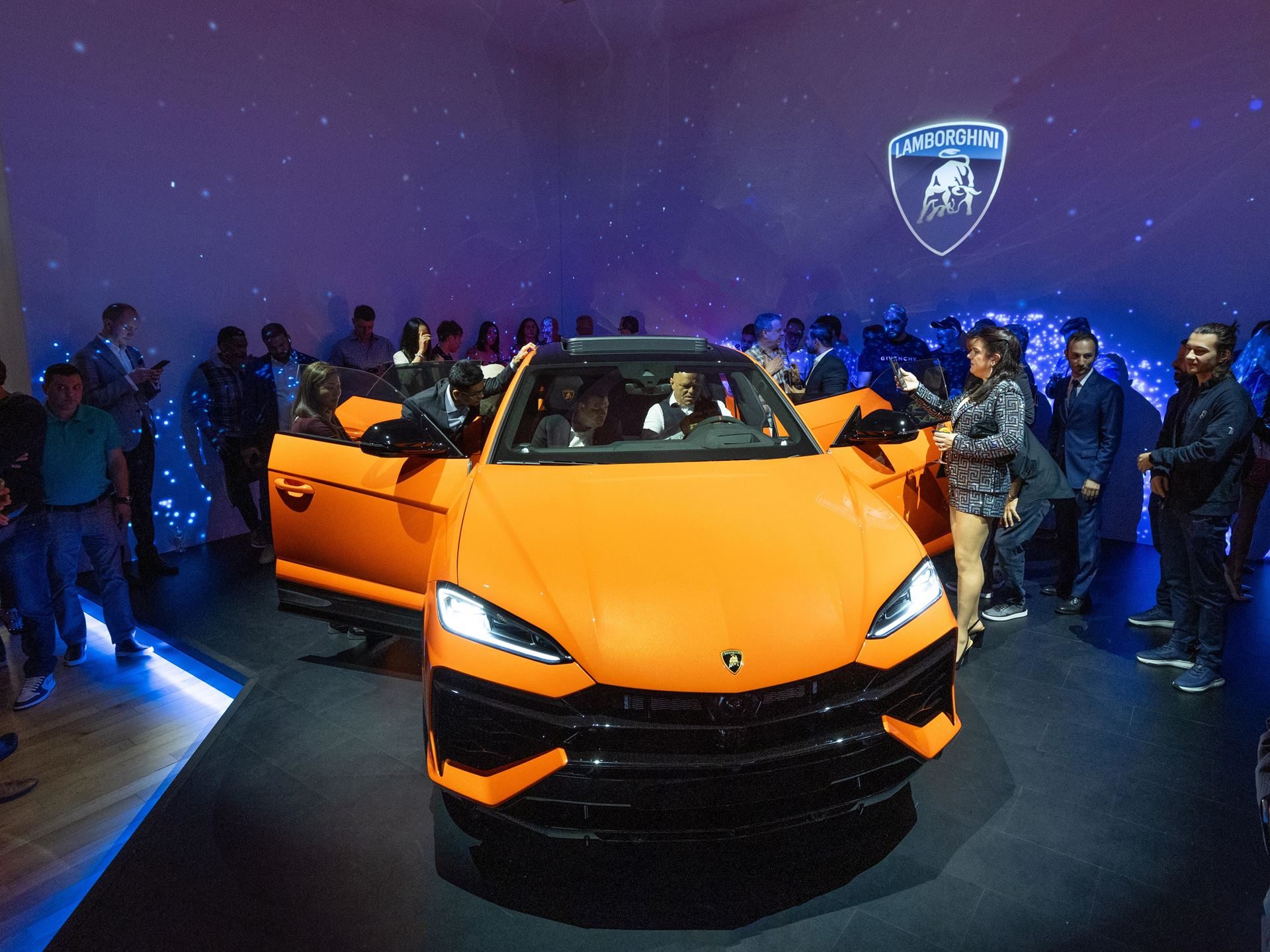 Lamborghini Brings The 789-Hp Urus Se To New York, Betting Its Best-Seller On Hybrid Power 9 Attendees gather around the orange lamborghini urus se at its us premiere, with some sitting inside the vehicle under a large lamborghini logo