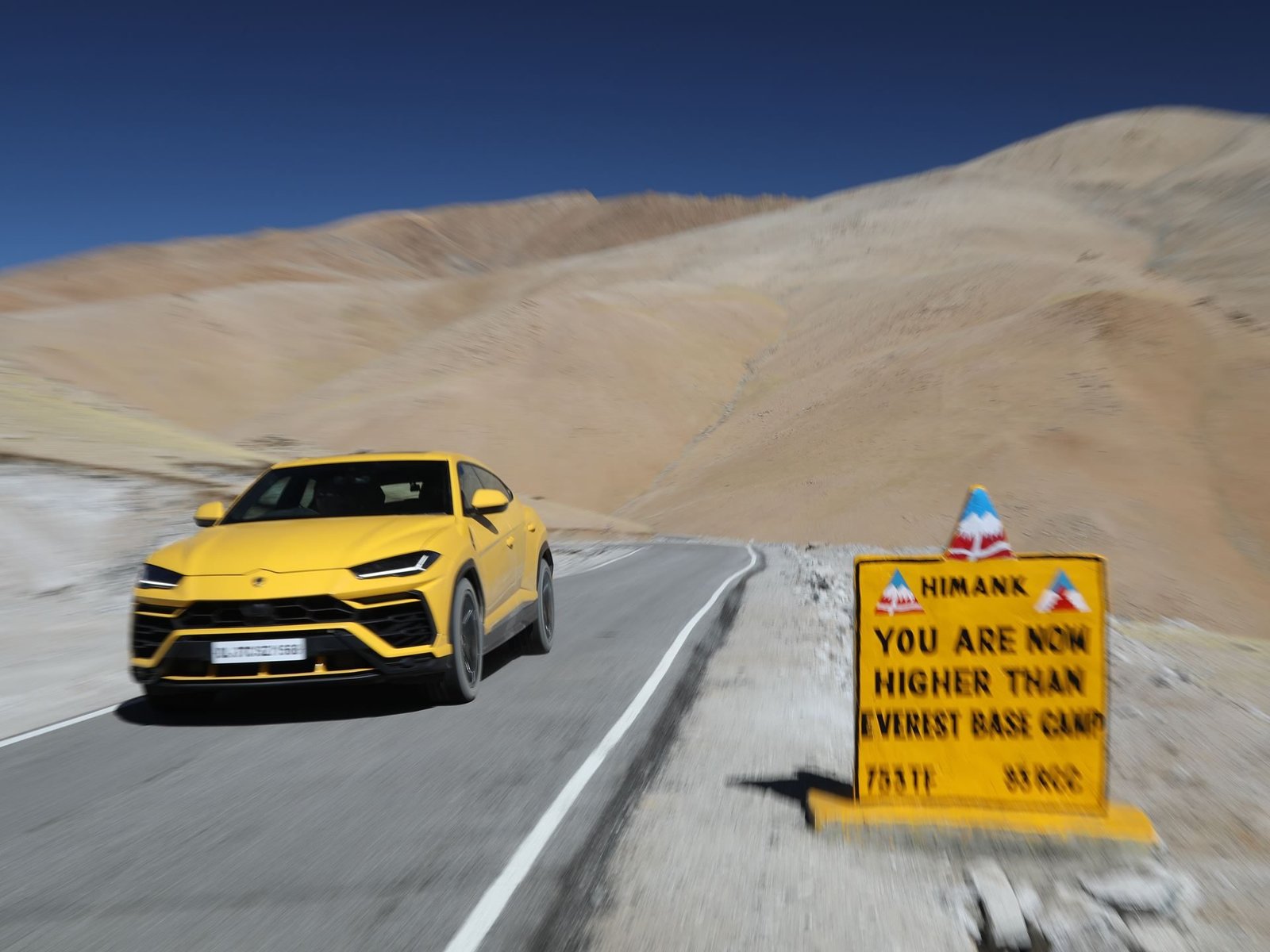 Yellow Lamborghini Urus driving on a winding mountain road past a sign reading Higher Than Everest Base Camp