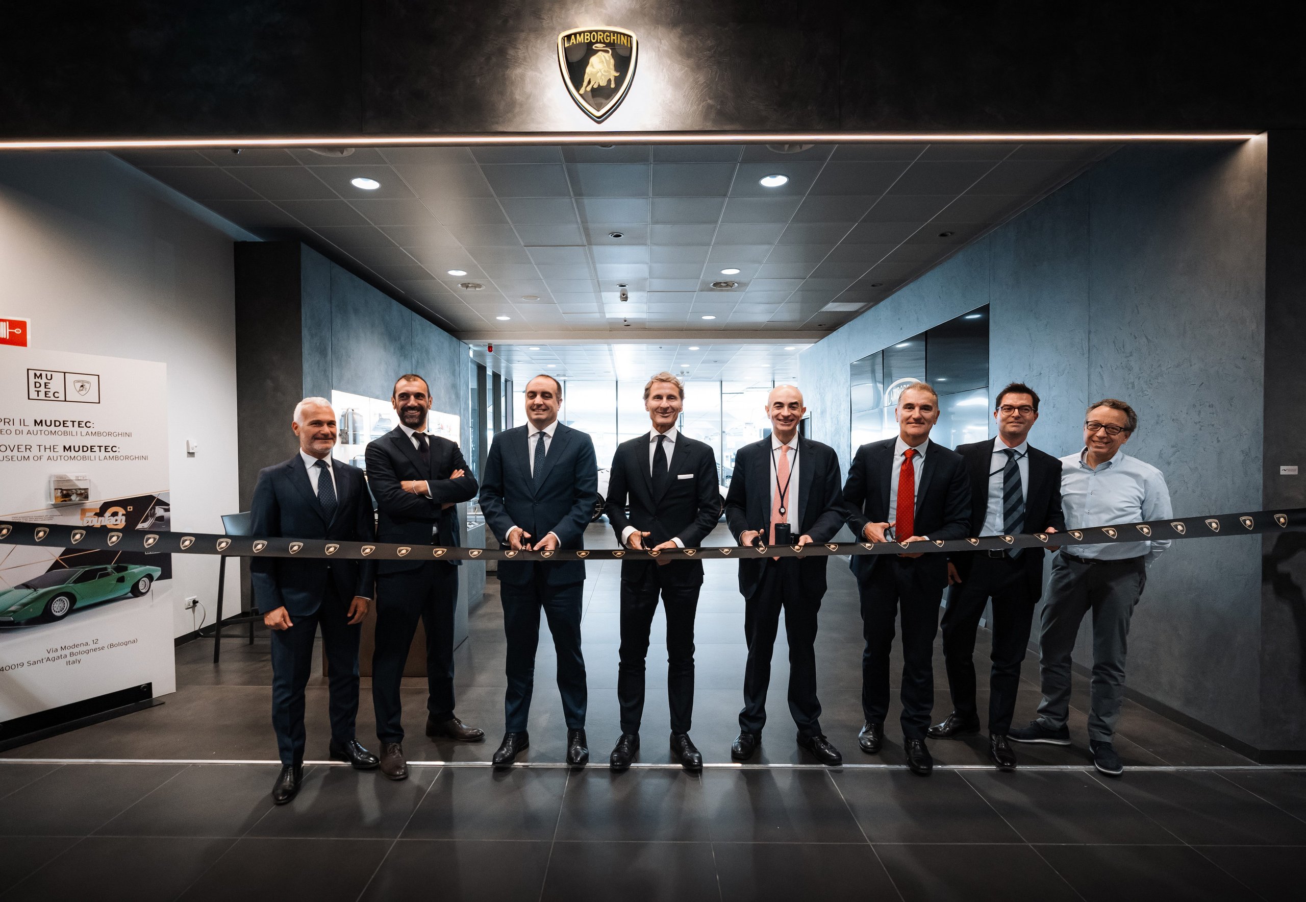 Lamborghini Turns Bologna Airport Into A Brand Experience, Complete With Urus Vip Shuttle And Huracán Follow-Me Car 5 Eight executives holding a ribbon in front of a lamborghini shield logo at the partnership renewal ceremony inside bologna airport