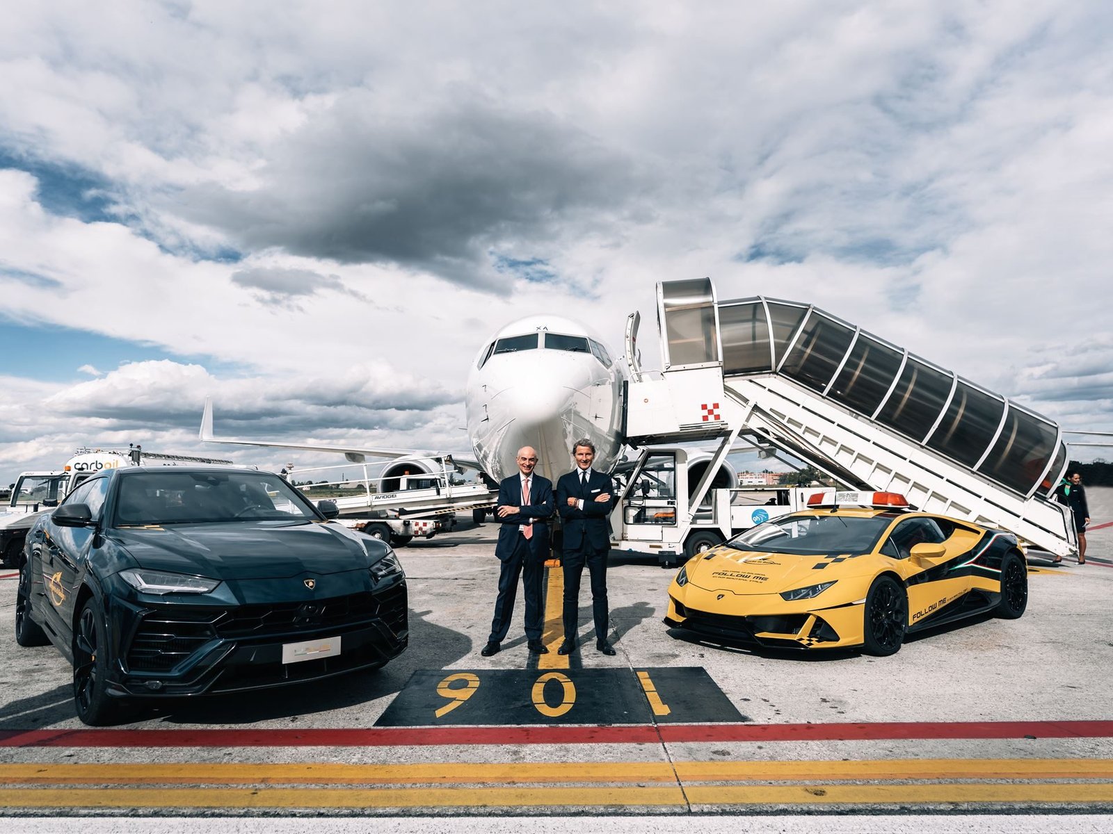 A black Lamborghini Urus and yellow Huracán follow-me car on the tarmac at Bologna Airport with a passenger jet in the background