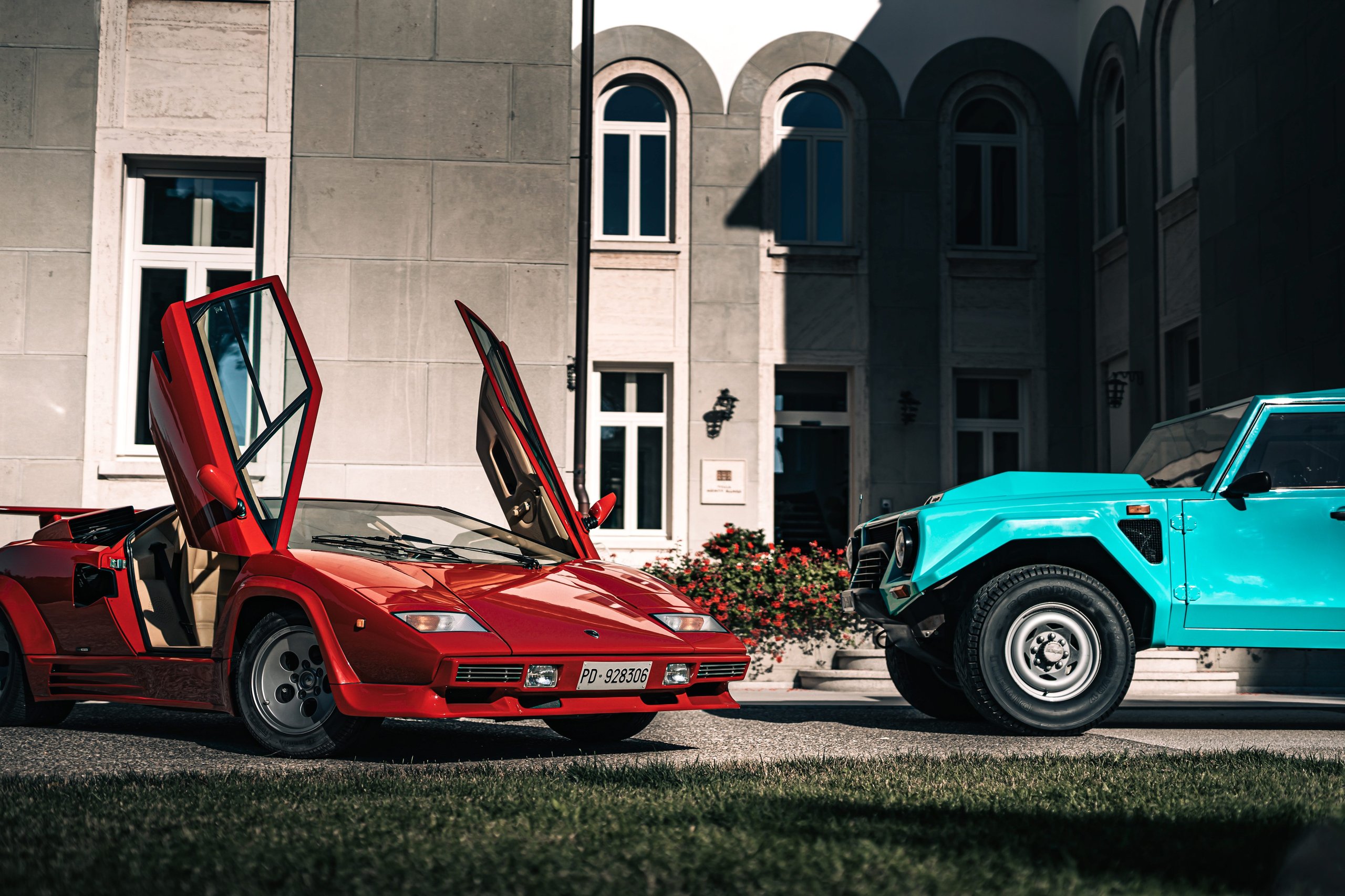 One V12, Two Wildly Different Lamborghinis: How The Countach And Lm 002 Proved The Twelve-Cylinder Could Do Anything 3 Red lamborghini countach with scissor doors open parked next to a teal lm 002 in front of a classic building