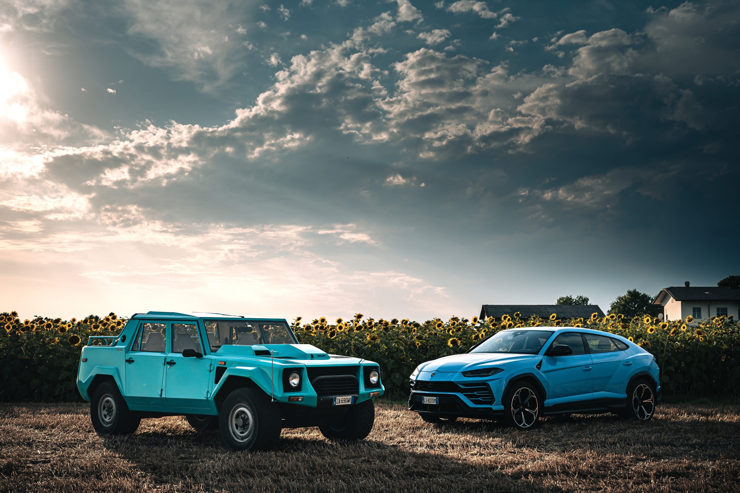One V12, Two Wildly Different Lamborghinis: How The Countach And Lm 002 Proved The Twelve-Cylinder Could Do Anything 9 Light blue lamborghini lm 002 and light blue lamborghini urus parked side by side in a sunflower field under dramatic sky