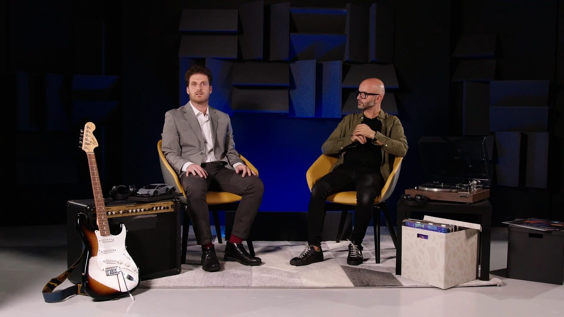 Alex trecarichi and mario mautone seated in a studio with sound panels, electric guitar, amplifier, and turntable