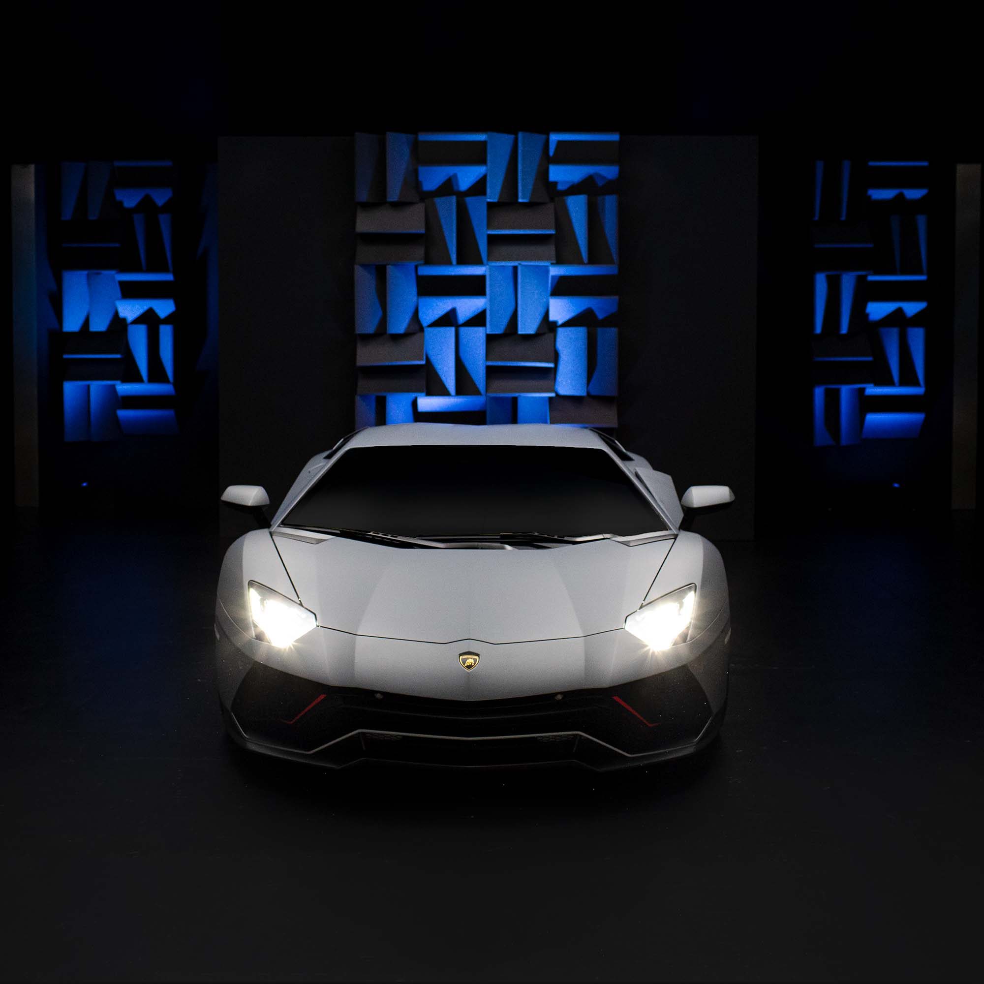 Grey lamborghini aventador front view with headlights illuminated in a dark studio with blue sound panels