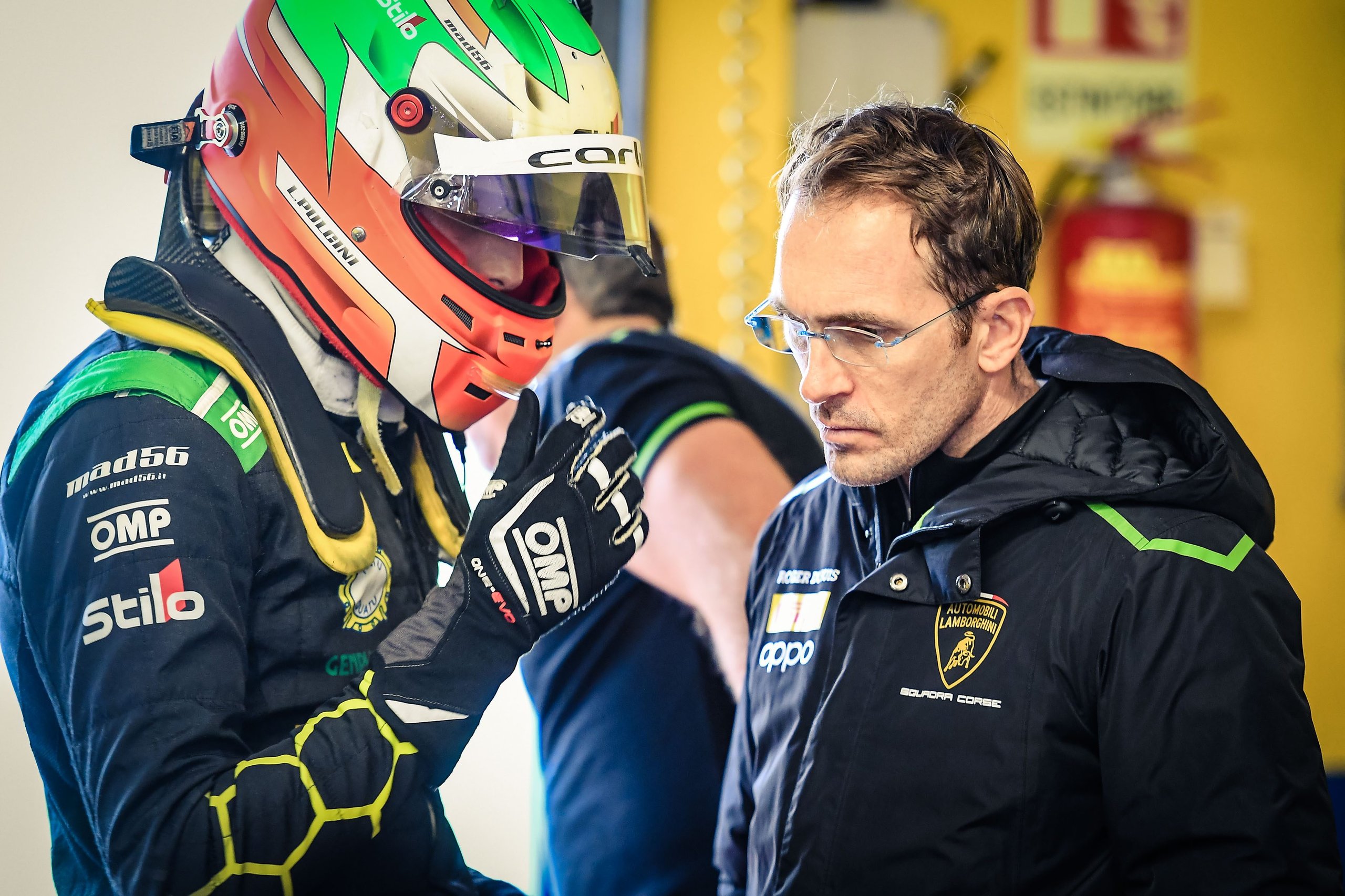 A lamborghini race driver in helmet and suit speaking with a team member wearing a squadra corse branded jacket in a pit garage