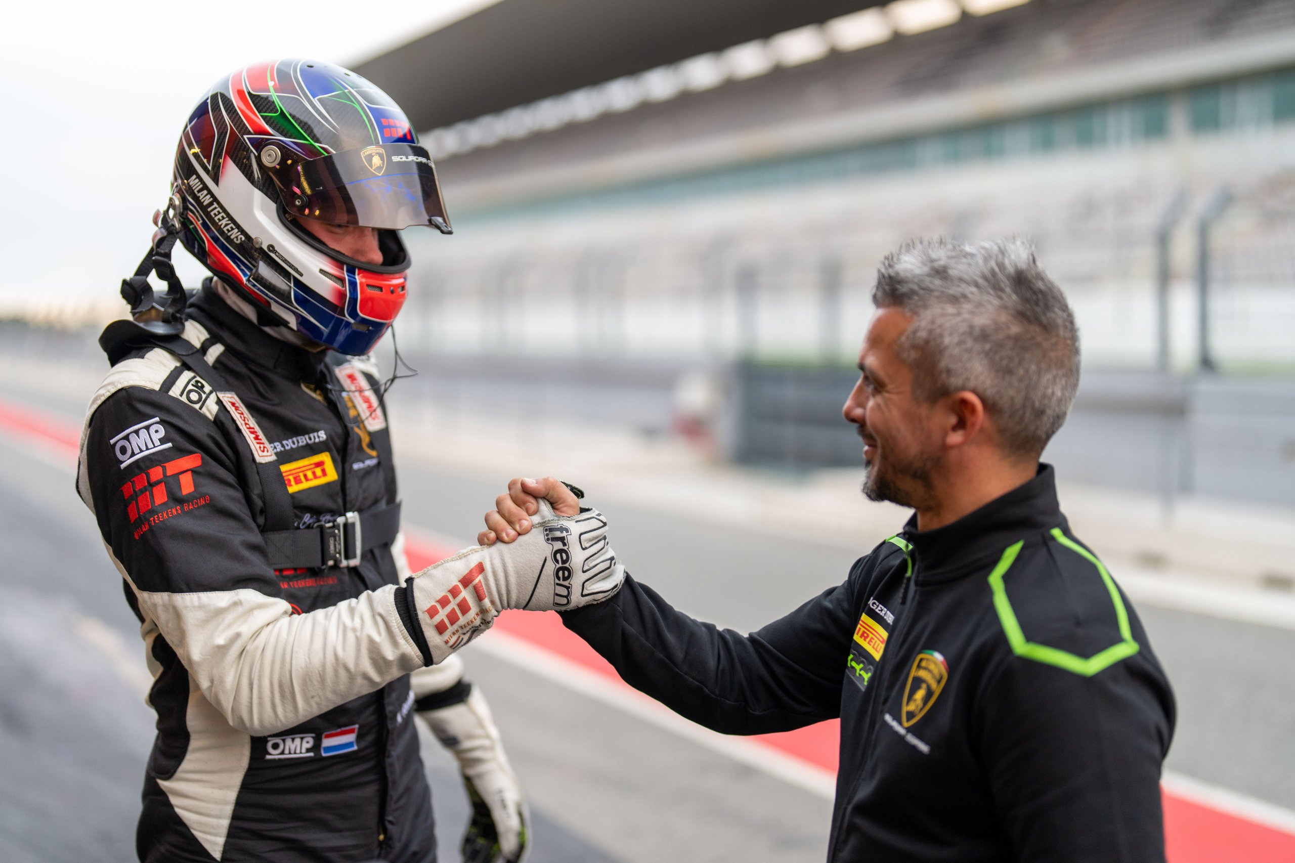 A lamborghini squadra corse team member in branded jacket shakes hands with a helmeted racing driver on a circuit