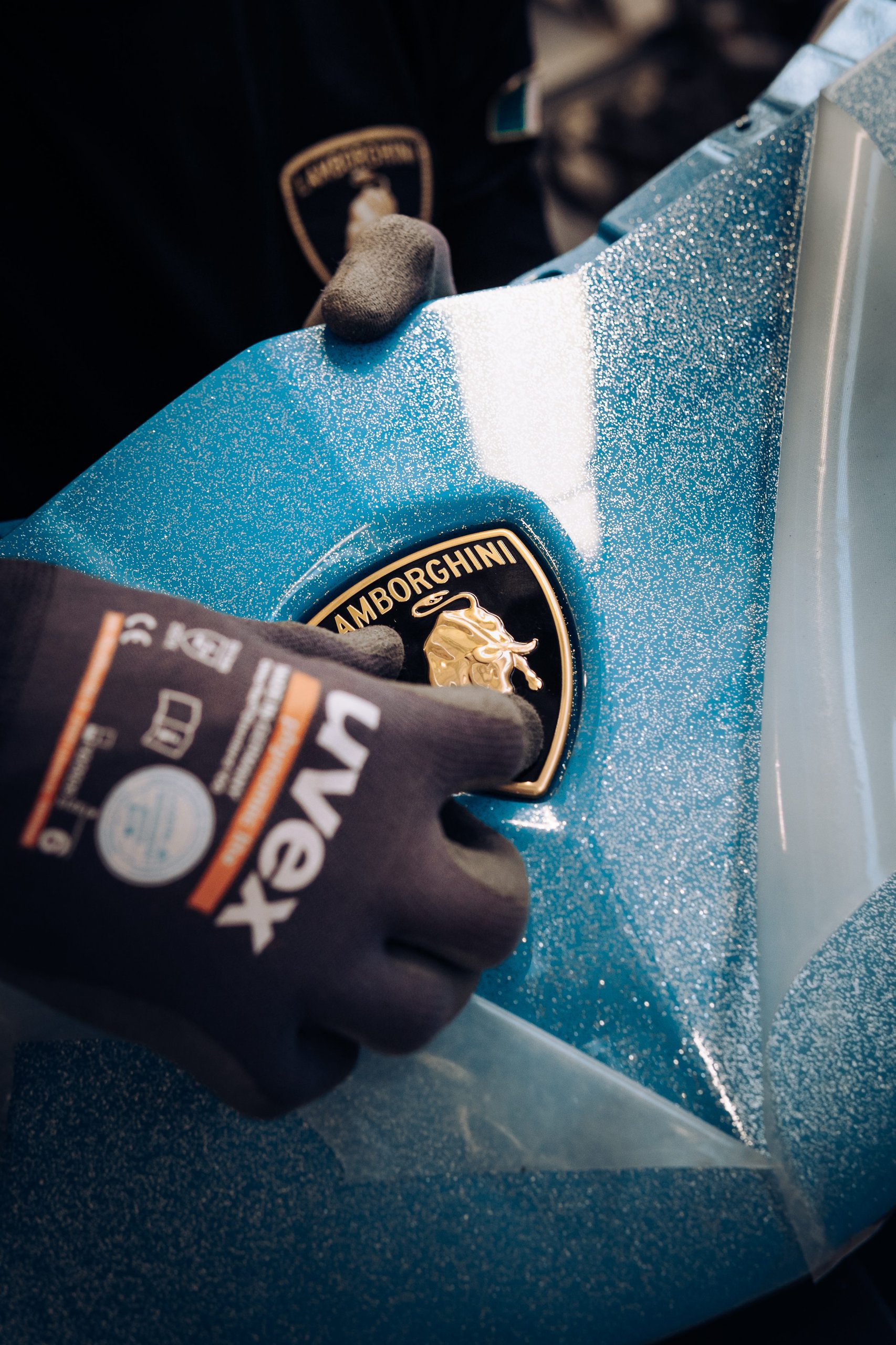 The Last Pure V12 Lamborghini Rolled Off The Line In Light Blue, And Nothing Will Replace What It Represented 13 Gloved hand applying the gold lamborghini emblem onto the sparkling blue body panel of the final aventador ultimae