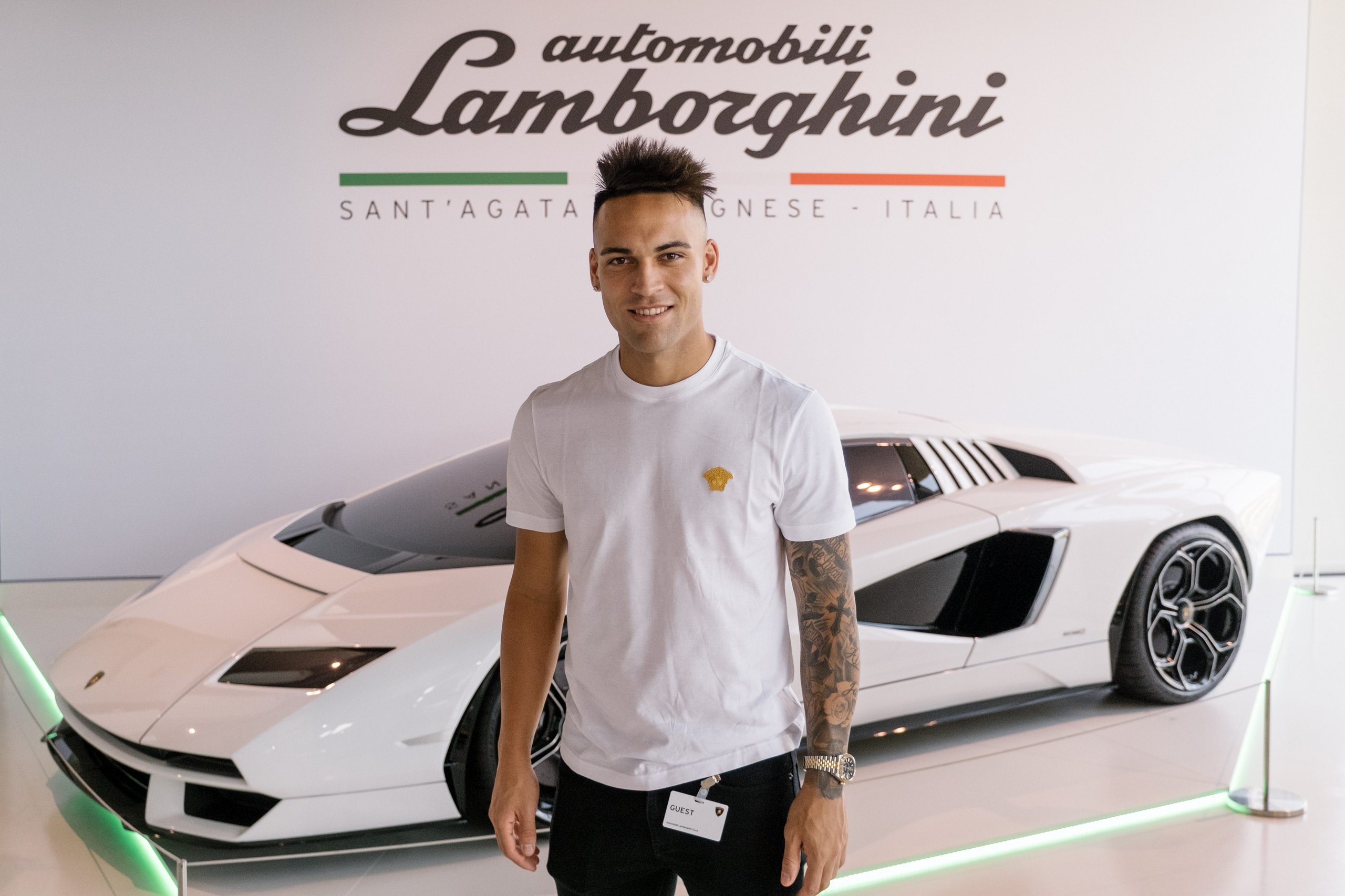 Lautaro martinez standing in the lamborghini showroom in front of a white countach lpi 800-4 with the automobili lamborghini logo on the wall behind him