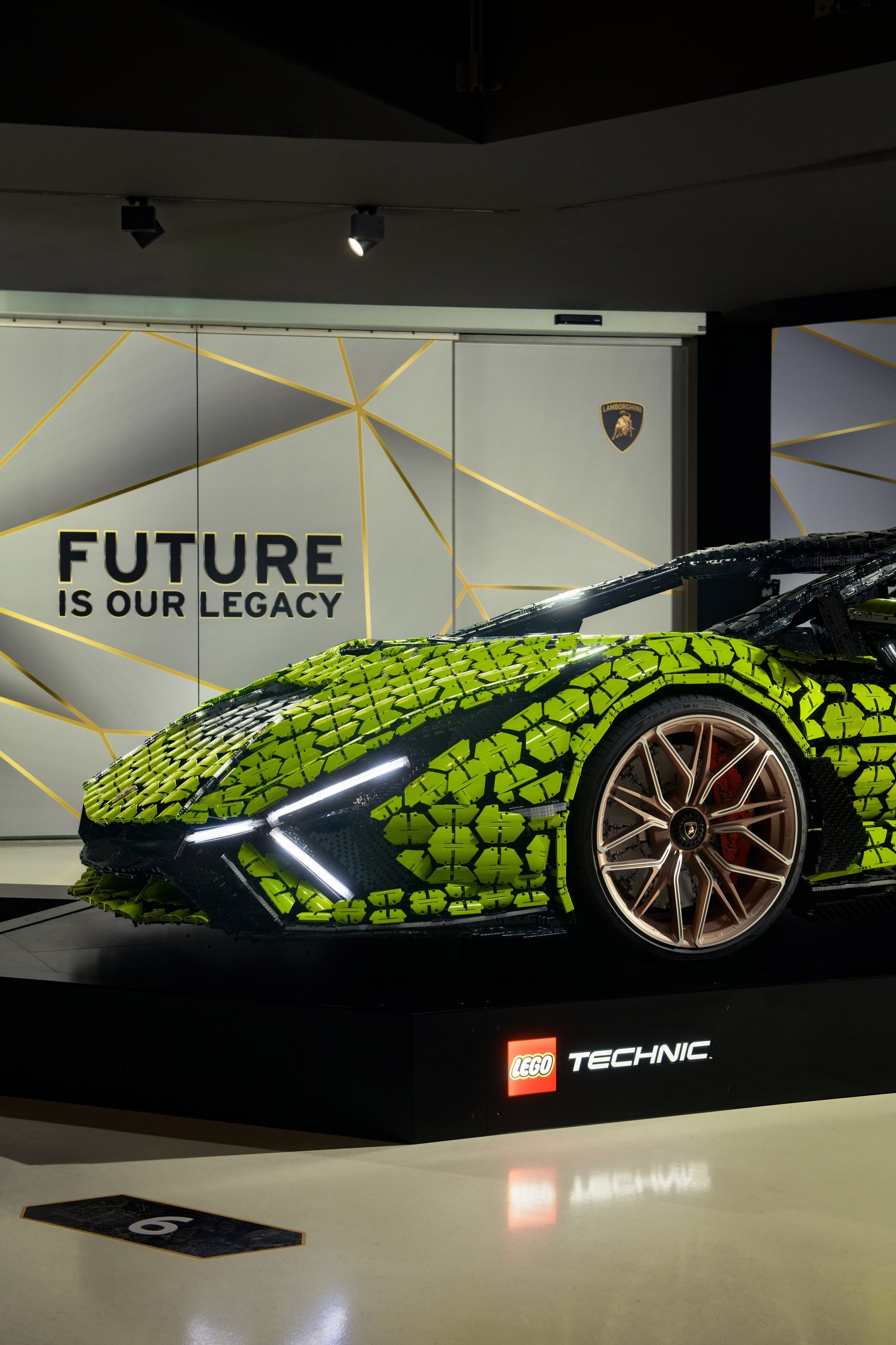 400,000 Bricks, One Sián: Inside The Life-Size Lego Technic Lamborghini That Got Painted In Sant'Agata 9 Side profile of the life-size lego technic lamborghini sián fkp 37 displayed on a black pedestal in front of a geometric 'future is our legacy' backdrop, showing y-shaped headlights and bronze wheels.