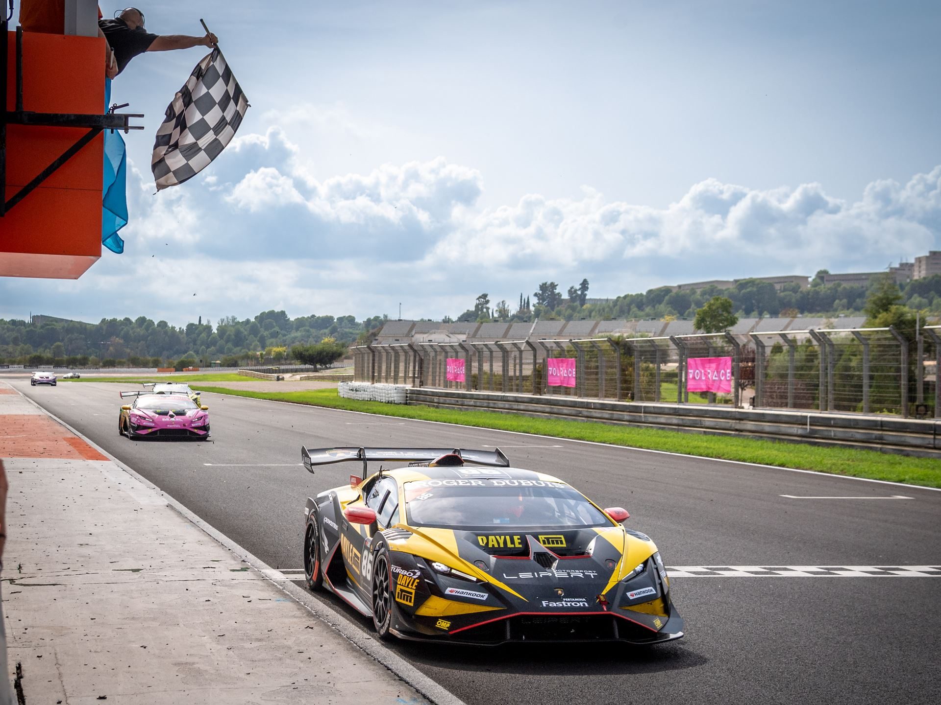 A yellow and black lamborghini huracán super trofeo evo2 crosses the finish line under a waving checkered flag in valencia