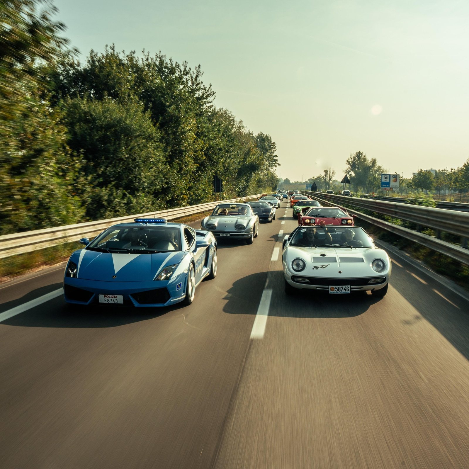 A blue Lamborghini Gallardo police car leads a convoy of classic Lamborghini models including a white Miura and silver 350 GT on a highway