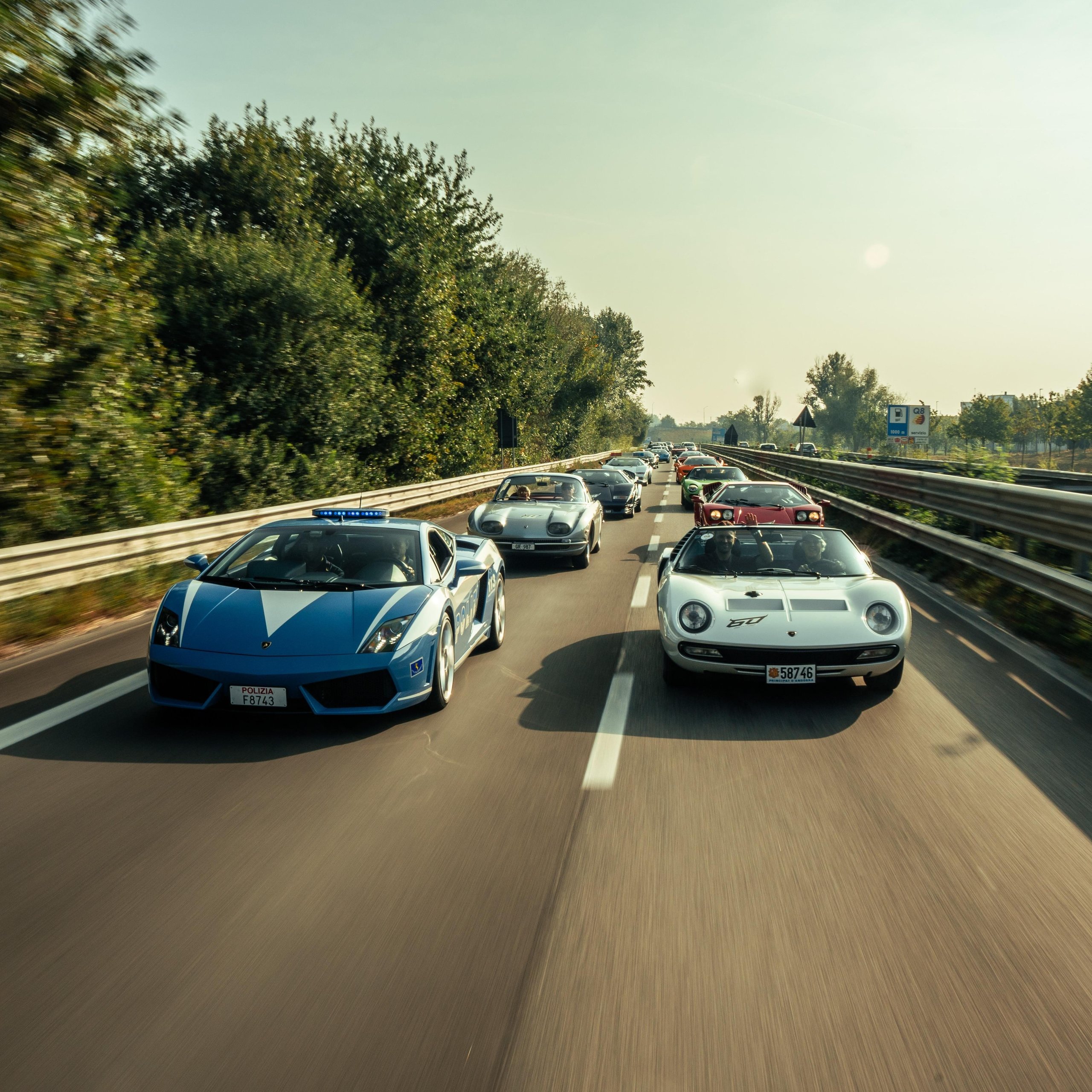 A blue lamborghini gallardo police car leads a convoy of classic lamborghini models including a white miura and silver 350 gt on a highway