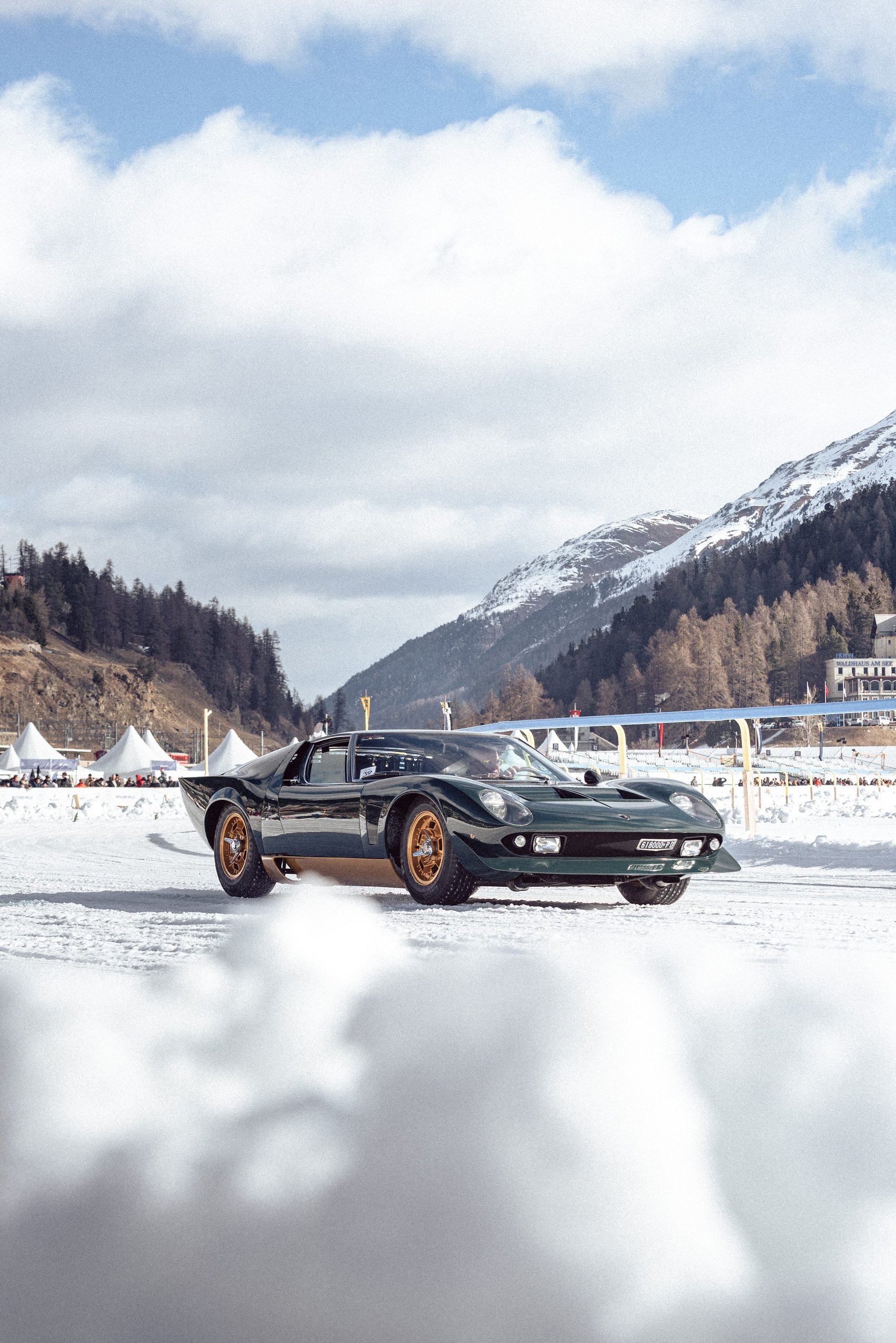 Dark green lamborghini miura millechiodi with gold wheels driving on a snowy track at st. Moritz with event tents and mountains