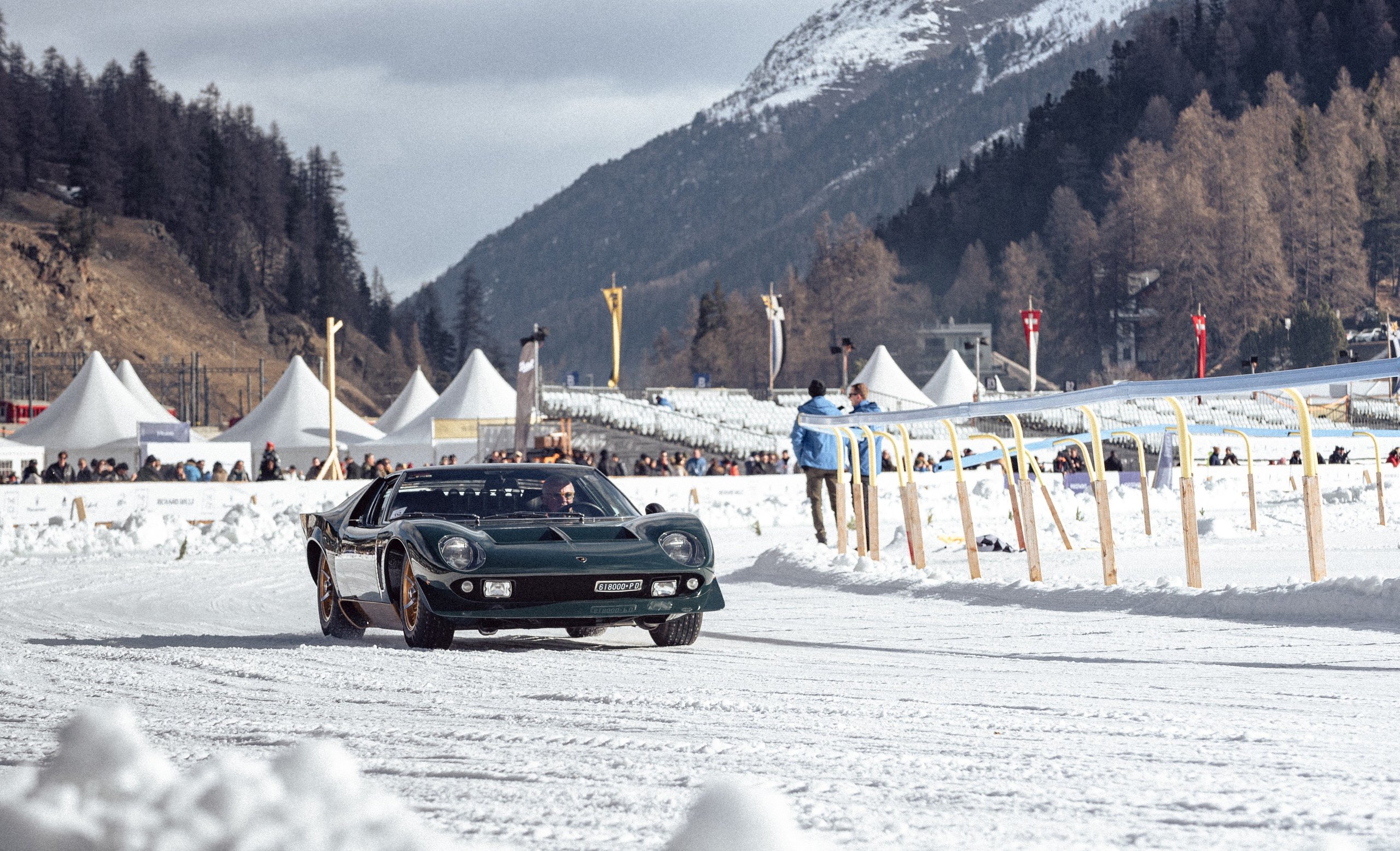 Dark green lamborghini miura p400 s millechiodi driving on a snowy track at the ice concours d'elegance in st. Moritz with spectators and mountains in the background